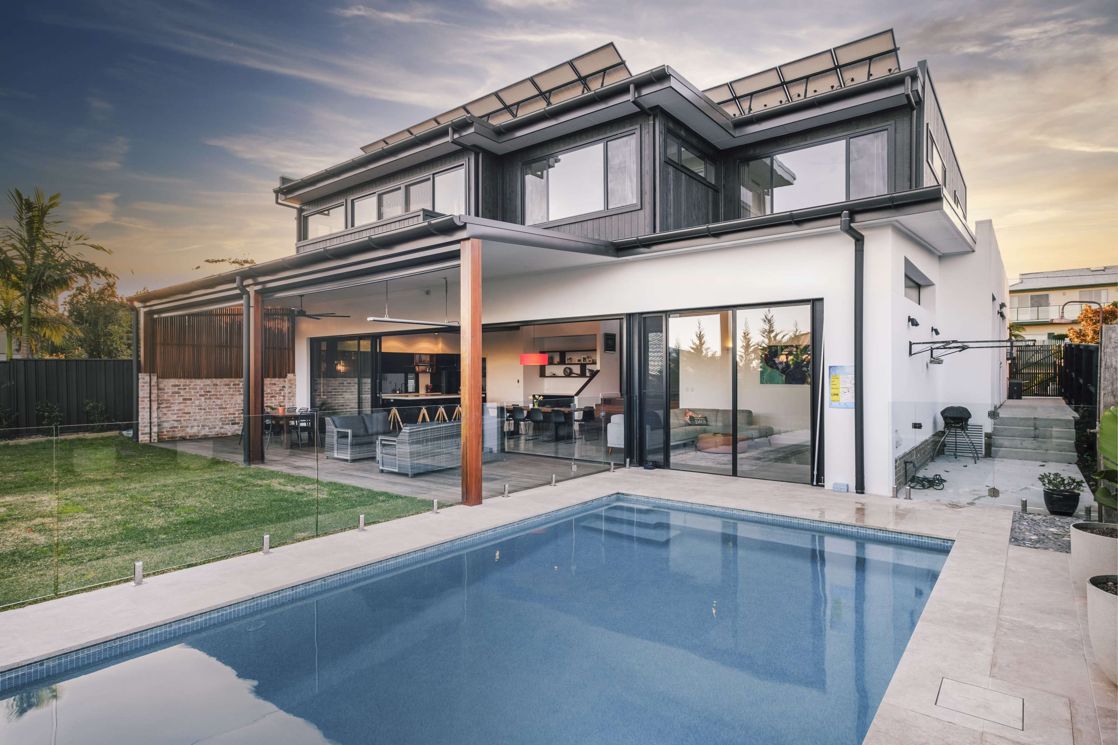 A modern two-story house with large windows and a swimming pool in the foreground, set against a colorful sunset.