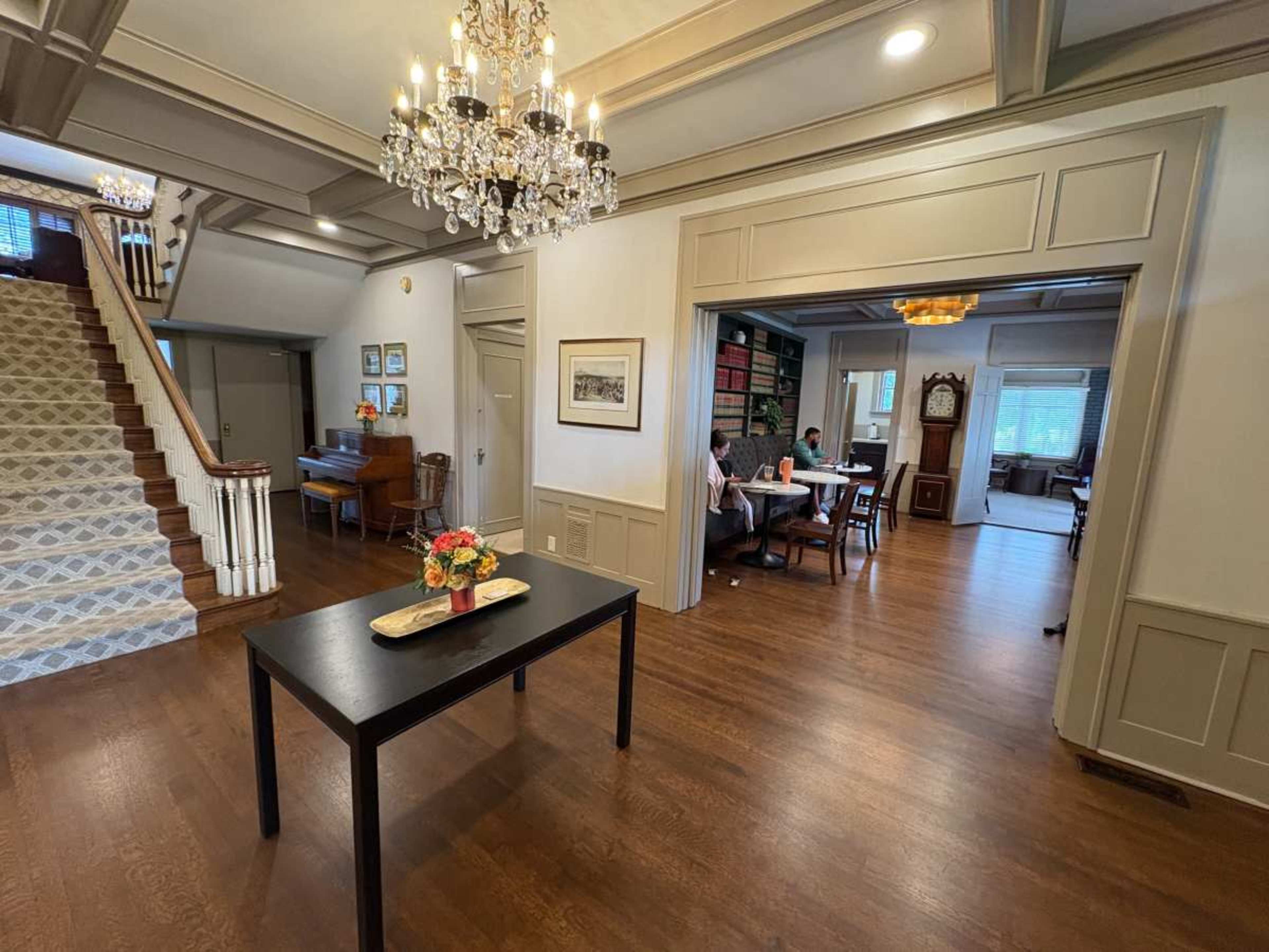 A well-lit foyer features a staircase, a table with flowers, and an open view into a dining area and living space where people are seated.