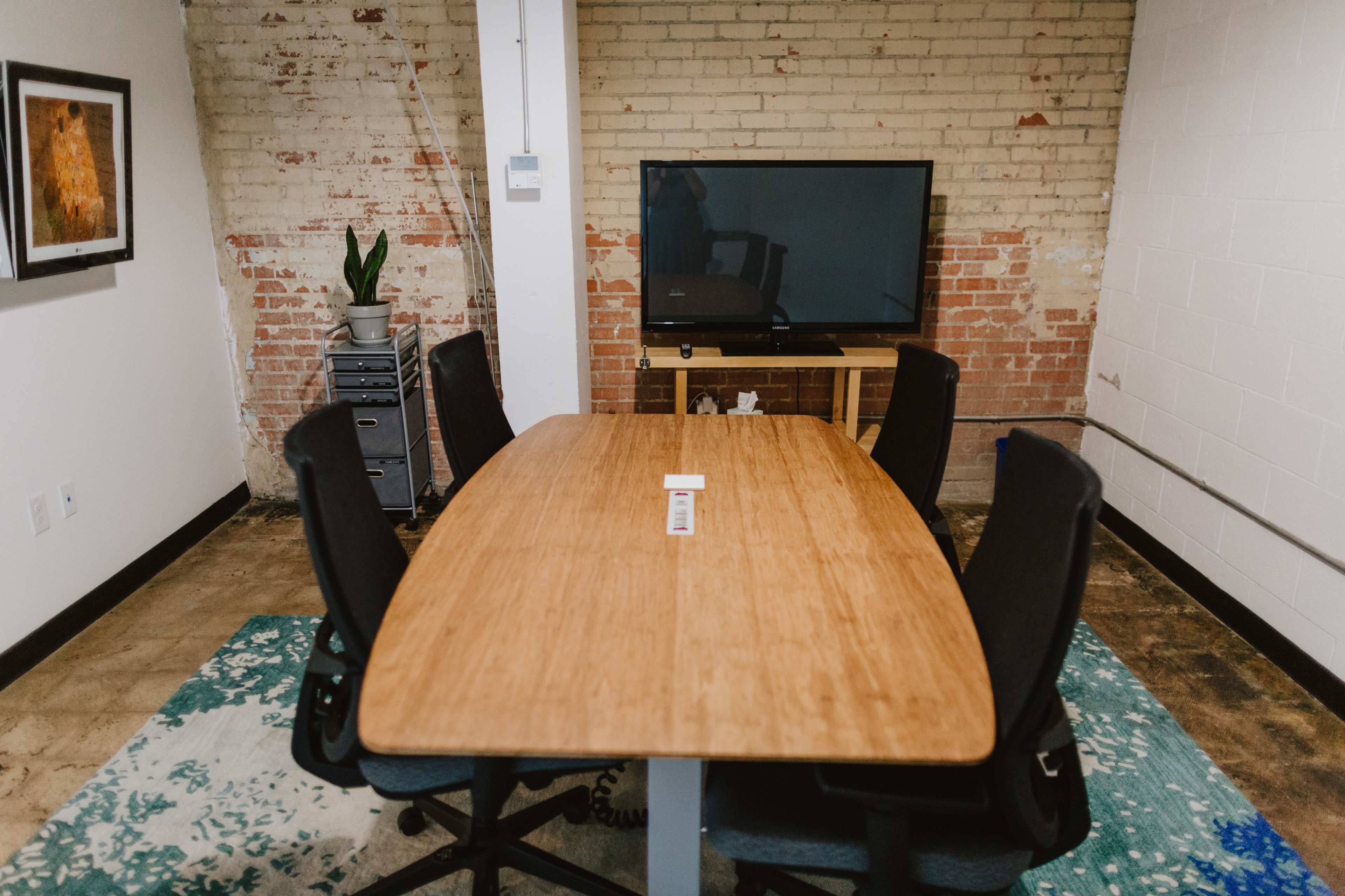 A conference room features a large wooden table surrounded by ergonomic chairs, with a television mounted on a wall and a potted plant nearby.