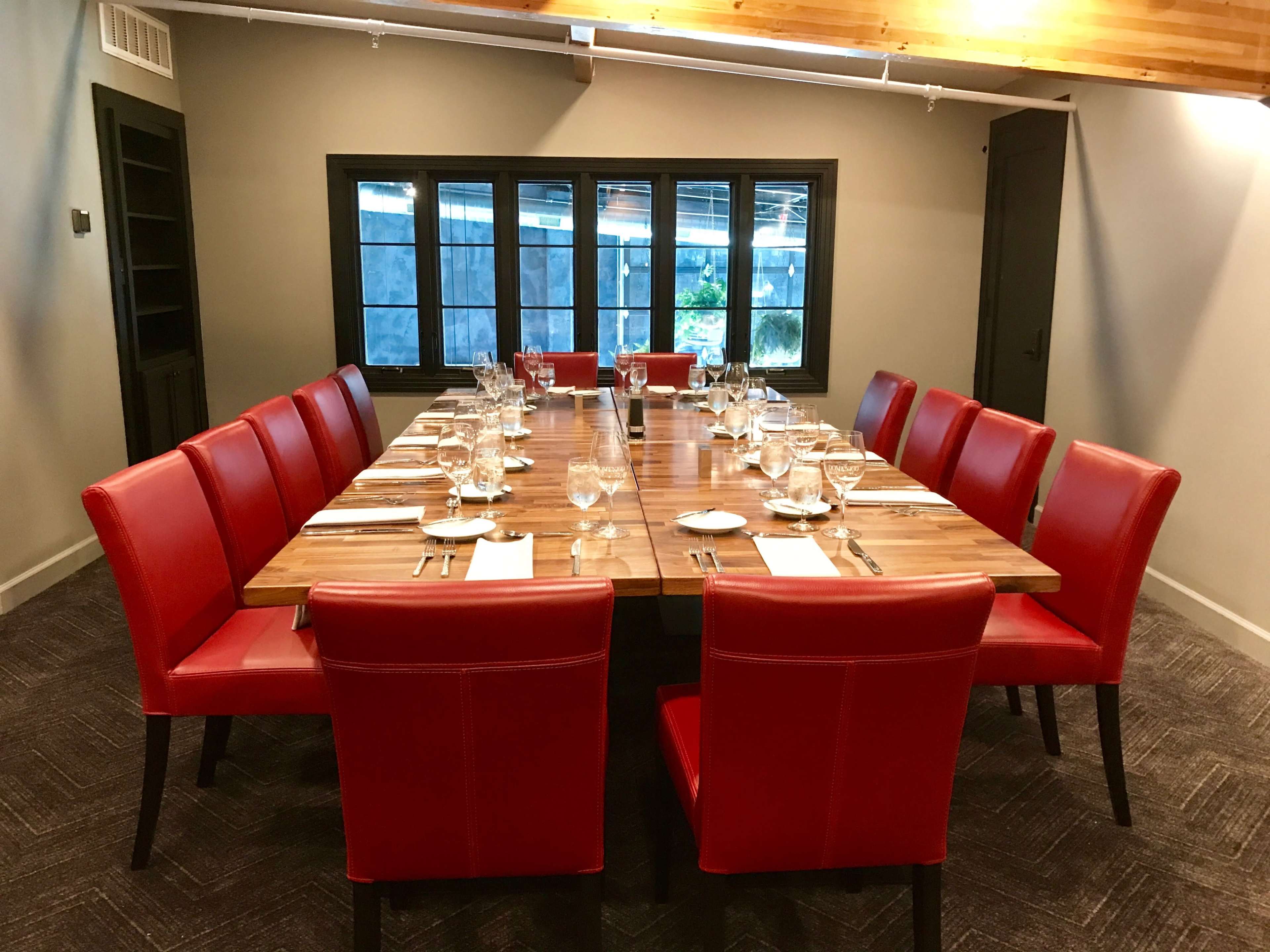 A long wooden dining table is set with glassware and silverware, surrounded by red chairs in a well-lit room with large windows.