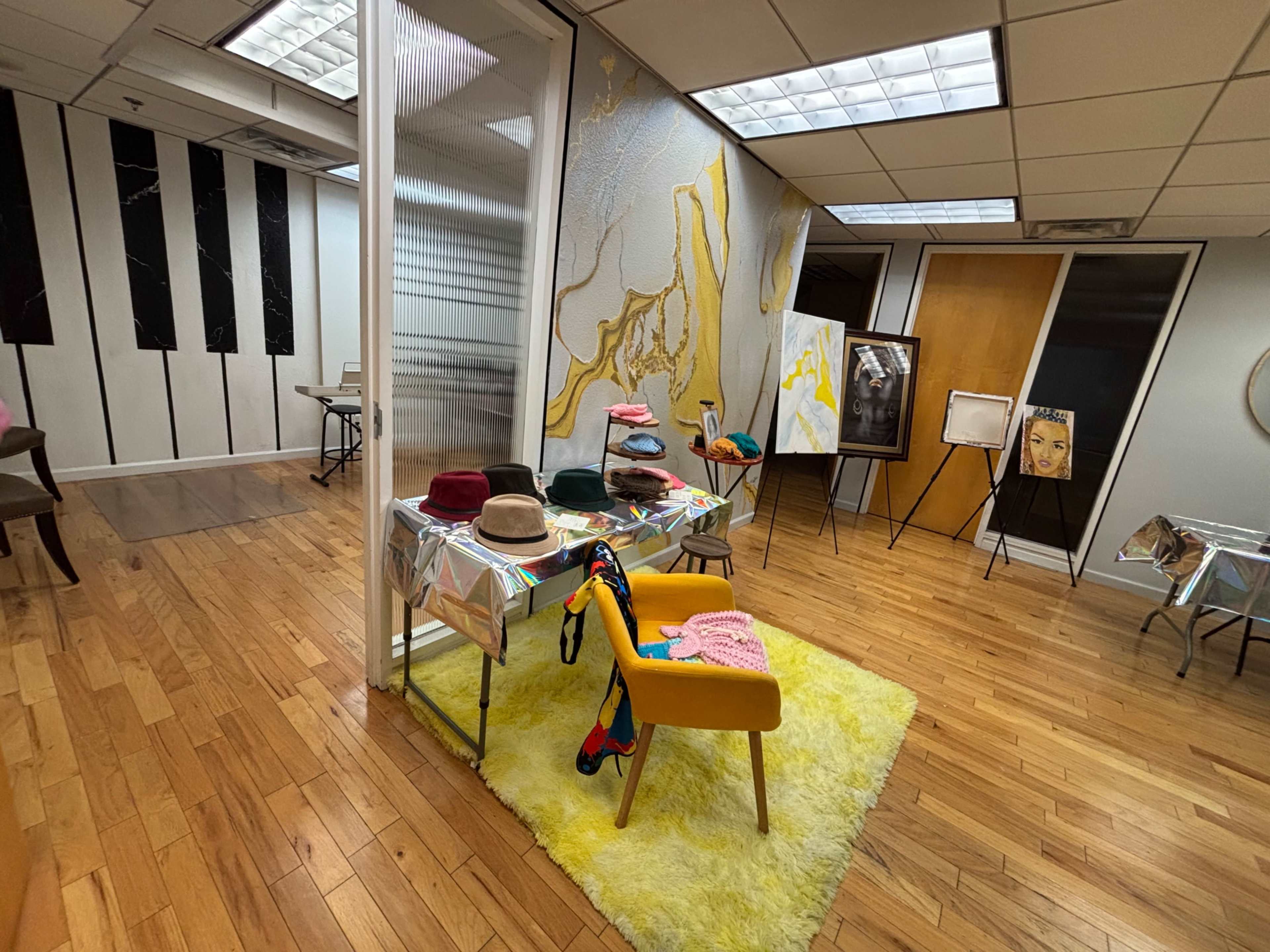 The image shows a spacious interior with wooden flooring, featuring a table adorned with various hats, a bright yellow rug, and artwork displayed on the walls.