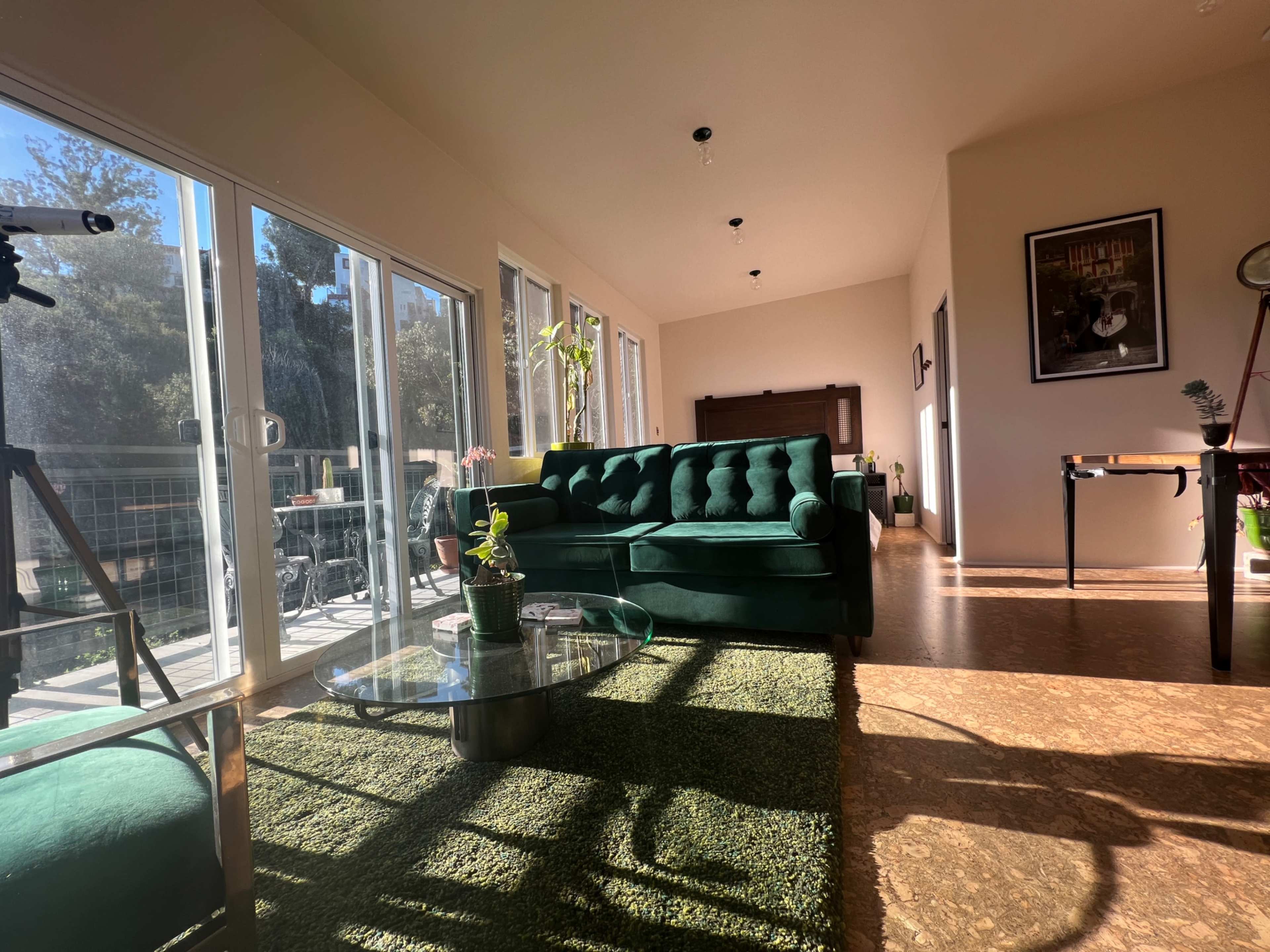 A well-lit living room featuring a green tufted sofa, a glass coffee table, and large windows displaying an outdoor view.