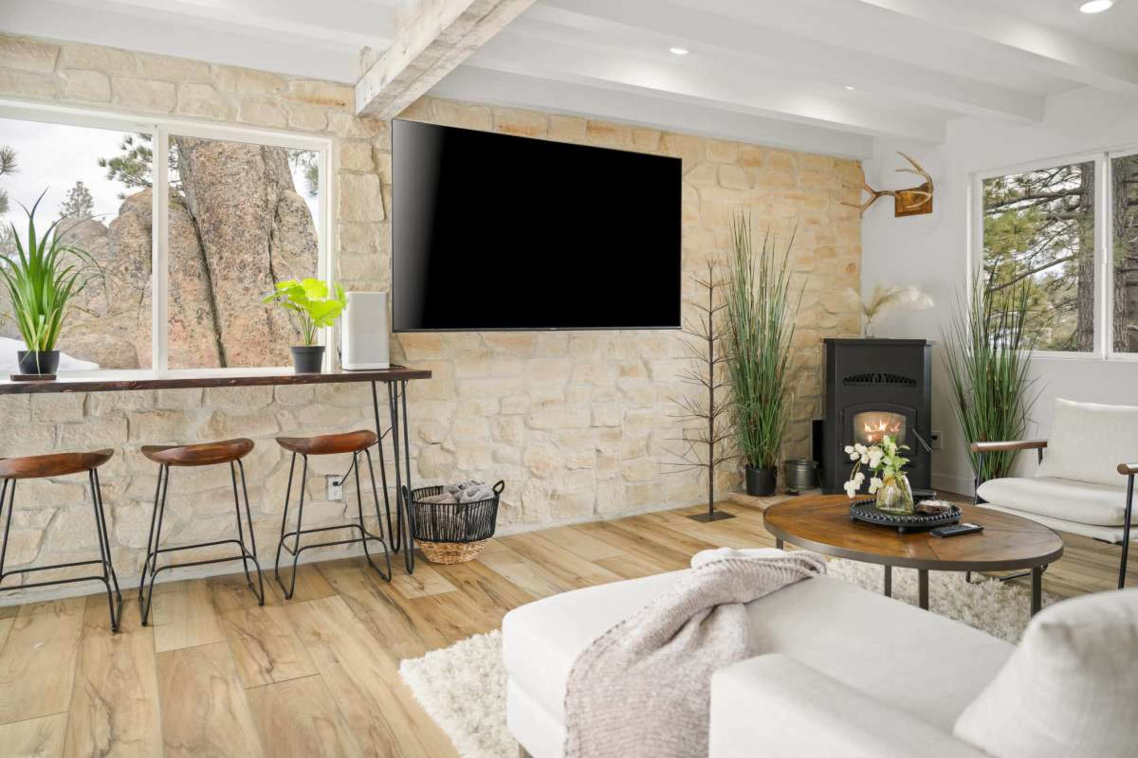 A modern living room features a stone wall, a large flat-screen TV, a round coffee table, and a small bar area with stools.