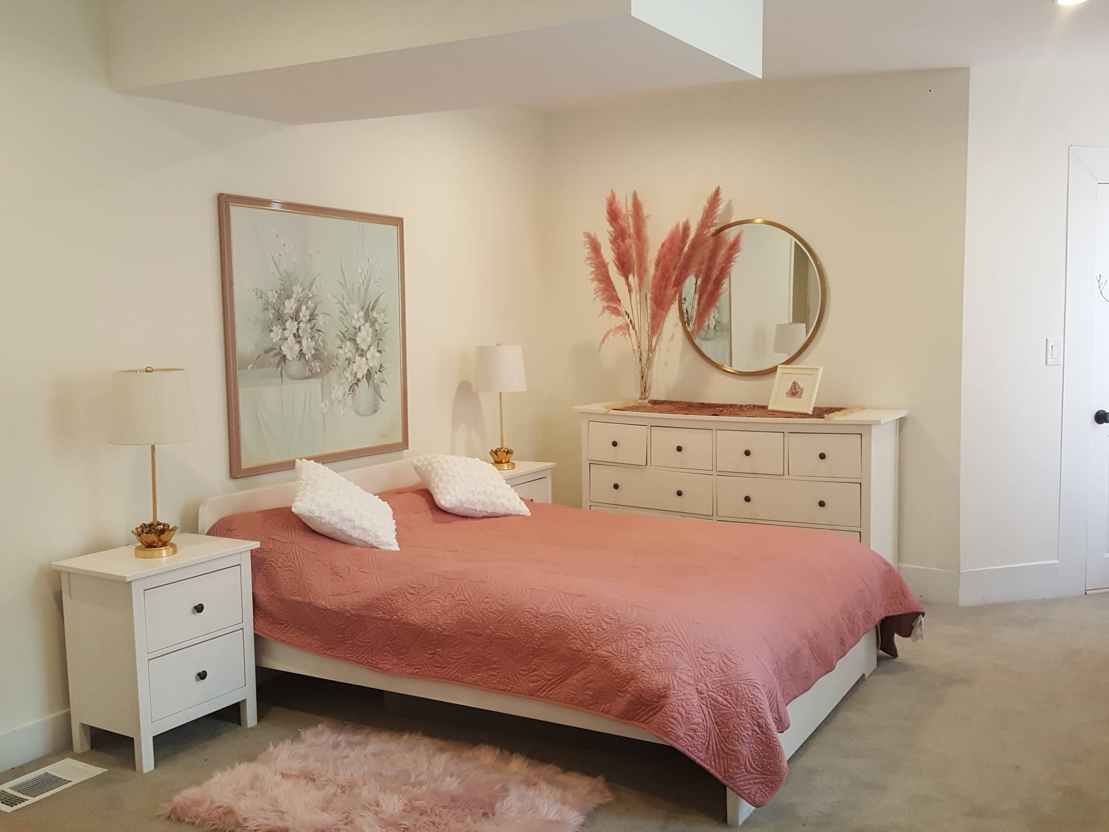 A neatly arranged bedroom features a bed with a pink quilt, bedside tables with lamps, a dresser, and decorative items, including a round mirror and pampas grass.