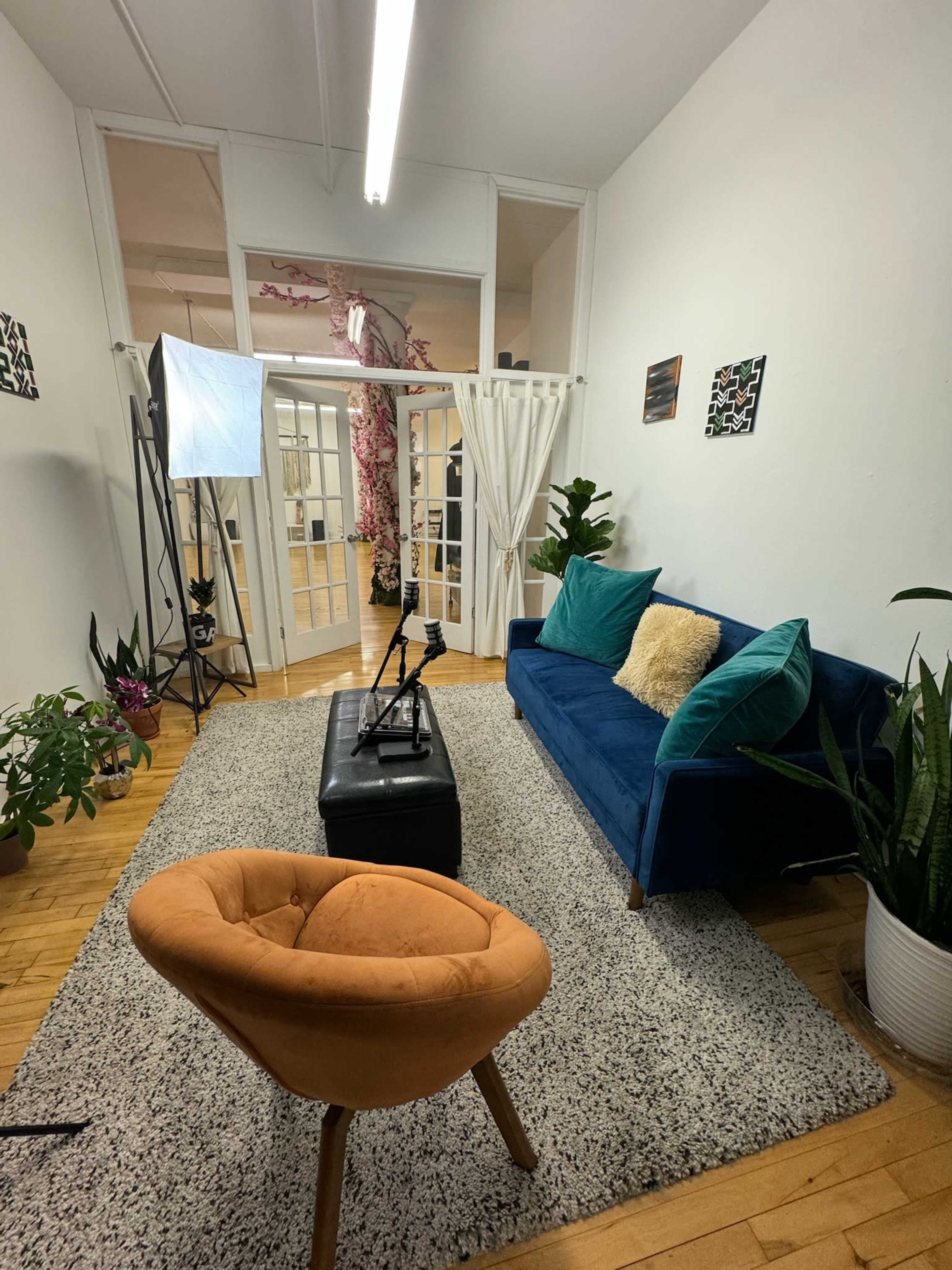 The room features a blue sofa with teal cushions, an orange chair, a black ottoman, and several plants, all arranged on a rug in a well-lit space with large double doors.