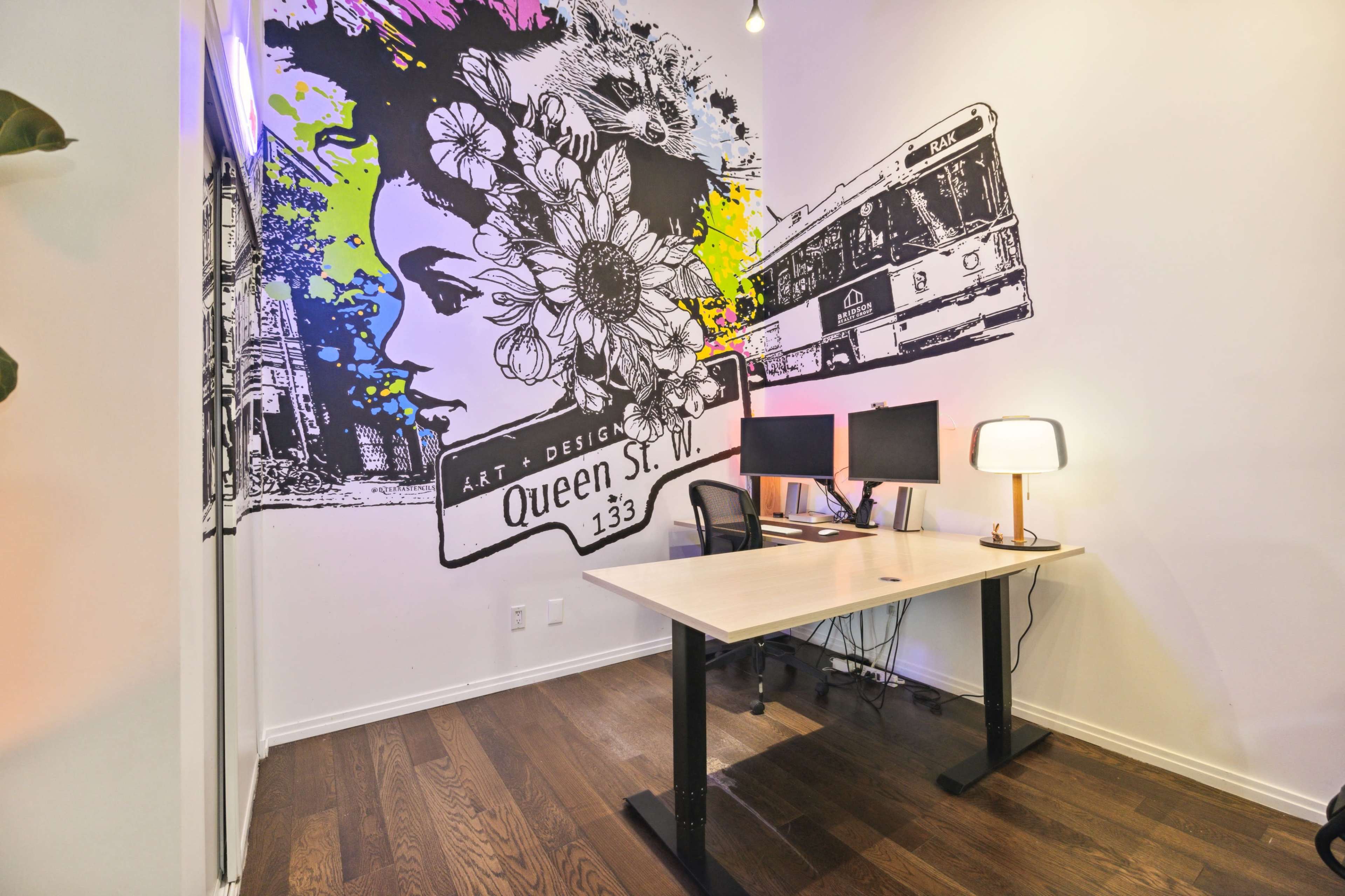 The image shows a modern office space featuring a desk, two computer monitors, and a large colorful wall mural depicting a woman's face and urban scenery.