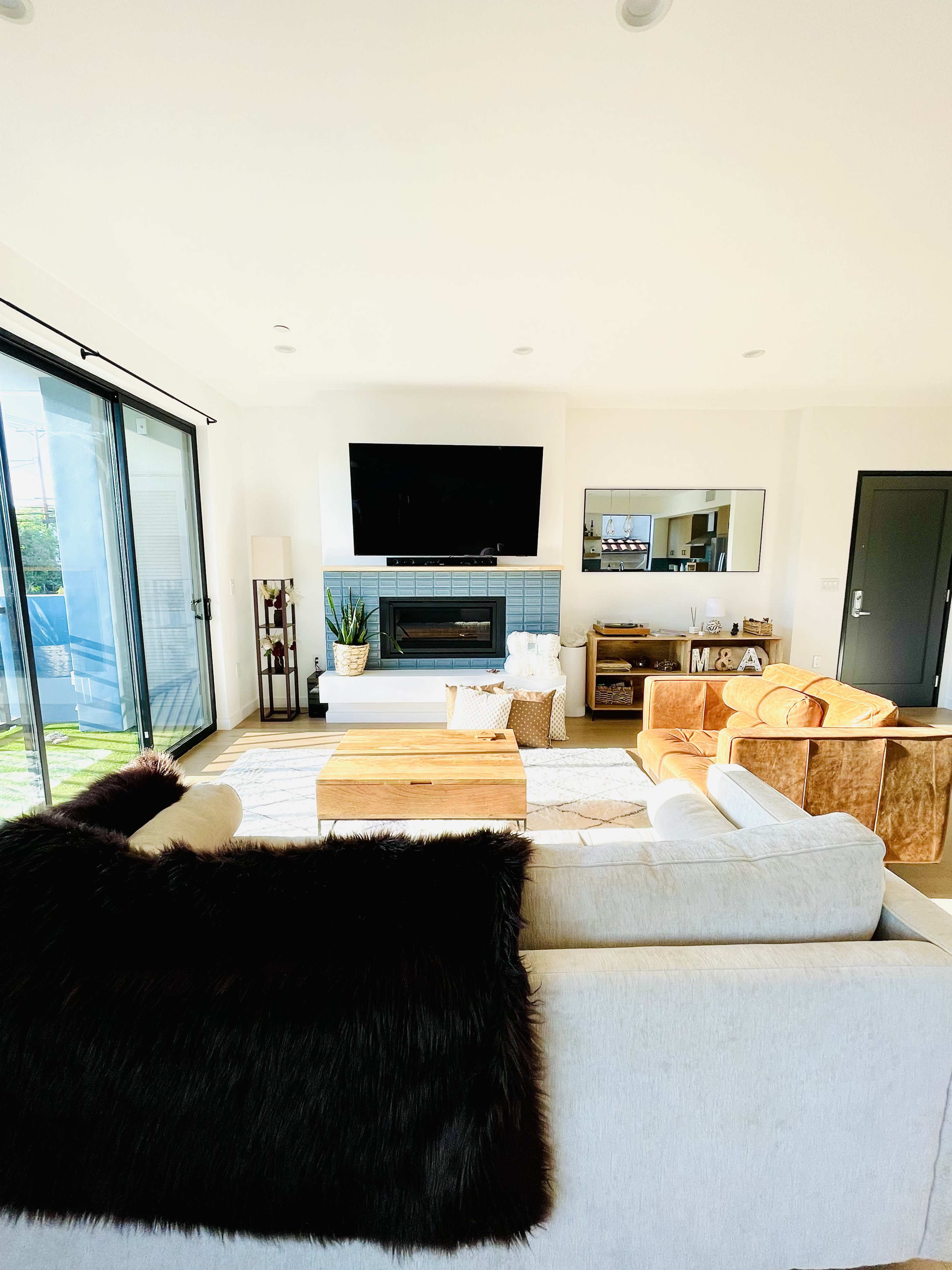 A modern living room features a television mounted above a fireplace, a wooden coffee table, and a mix of seating including a white sofa and an orange chair.