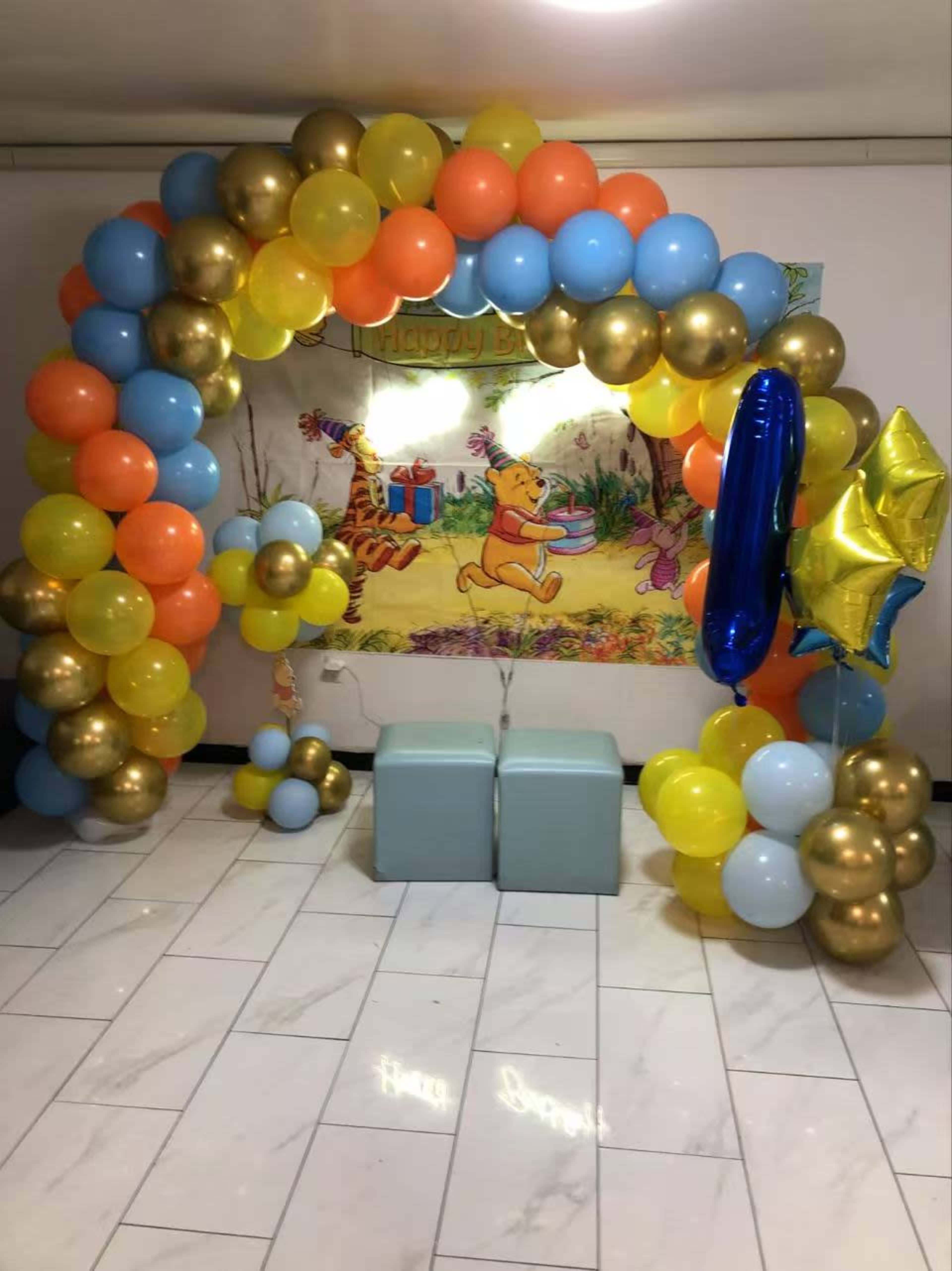A colorful balloon arch featuring blue, orange, yellow, and gold balloons frames a Winnie the Pooh birthday backdrop with two light blue chairs in front.