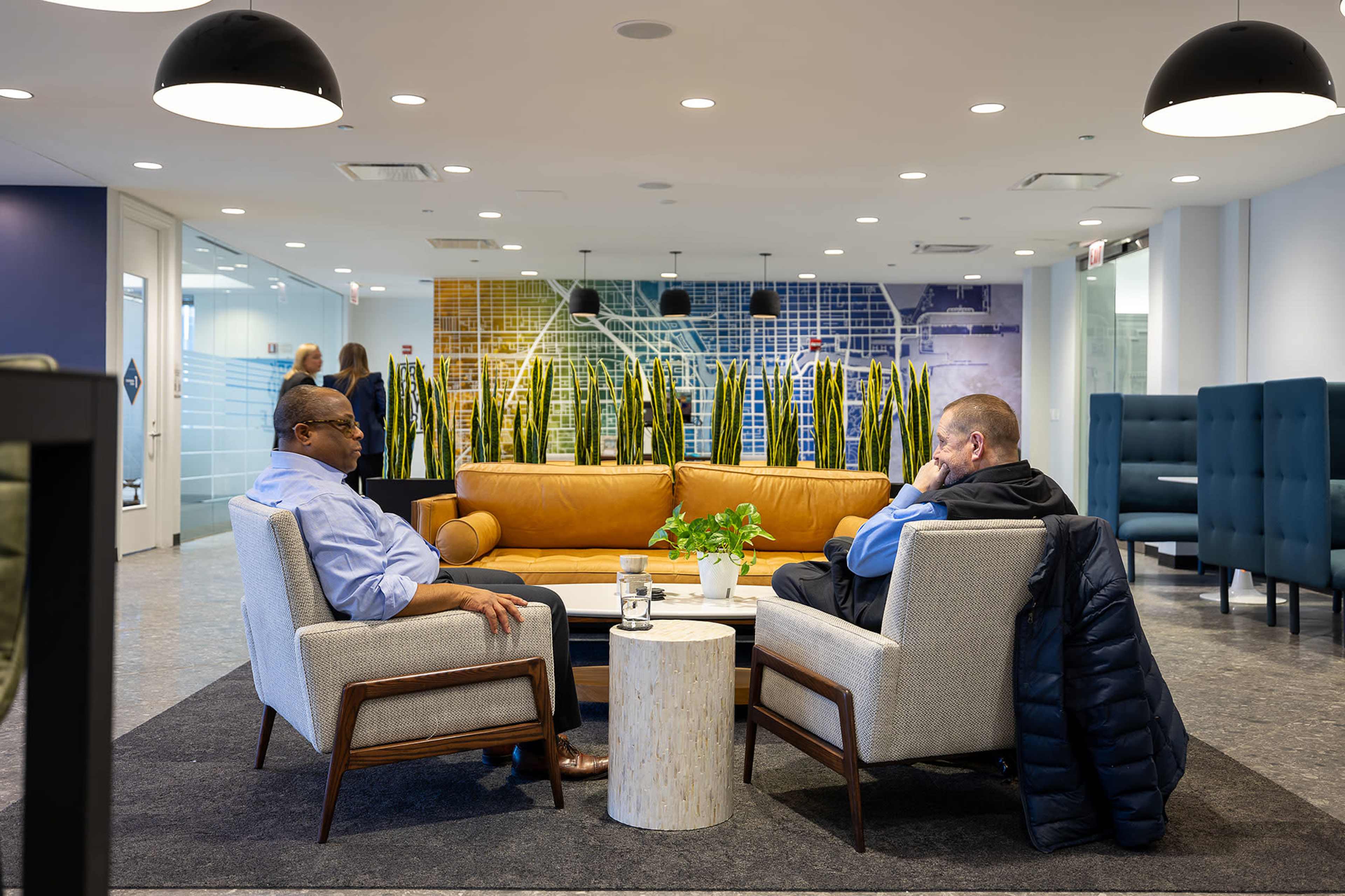 Two men are sitting in a modern office lounge area, engaged in conversation, with a sofa and decorative plants in the background.