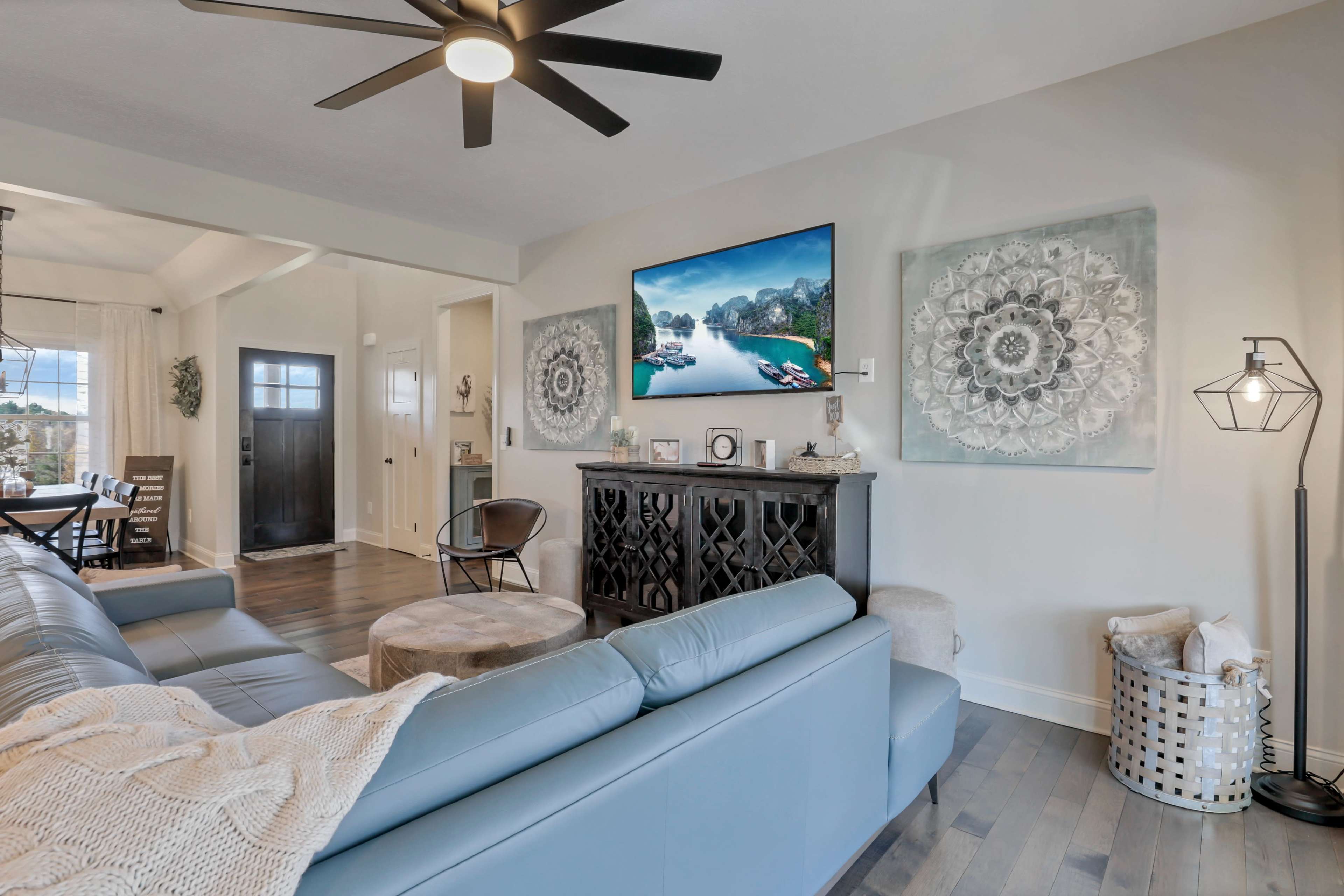 A modern living room features a light blue sectional couch, a wall-mounted television displaying a scenic landscape, and decorative wall art.