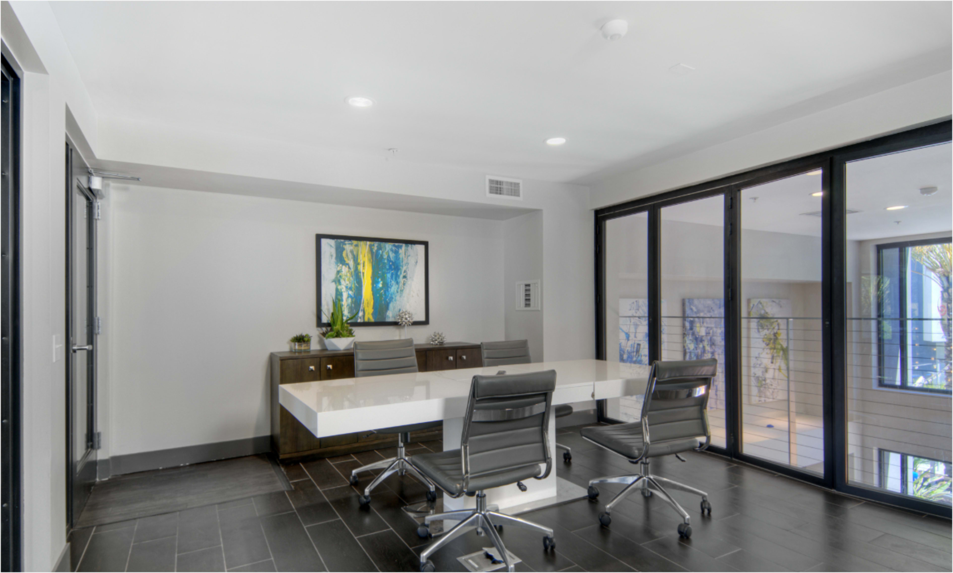 A modern conference room features a long white table surrounded by four black ergonomic chairs, with large glass doors leading to an outdoor balcony.