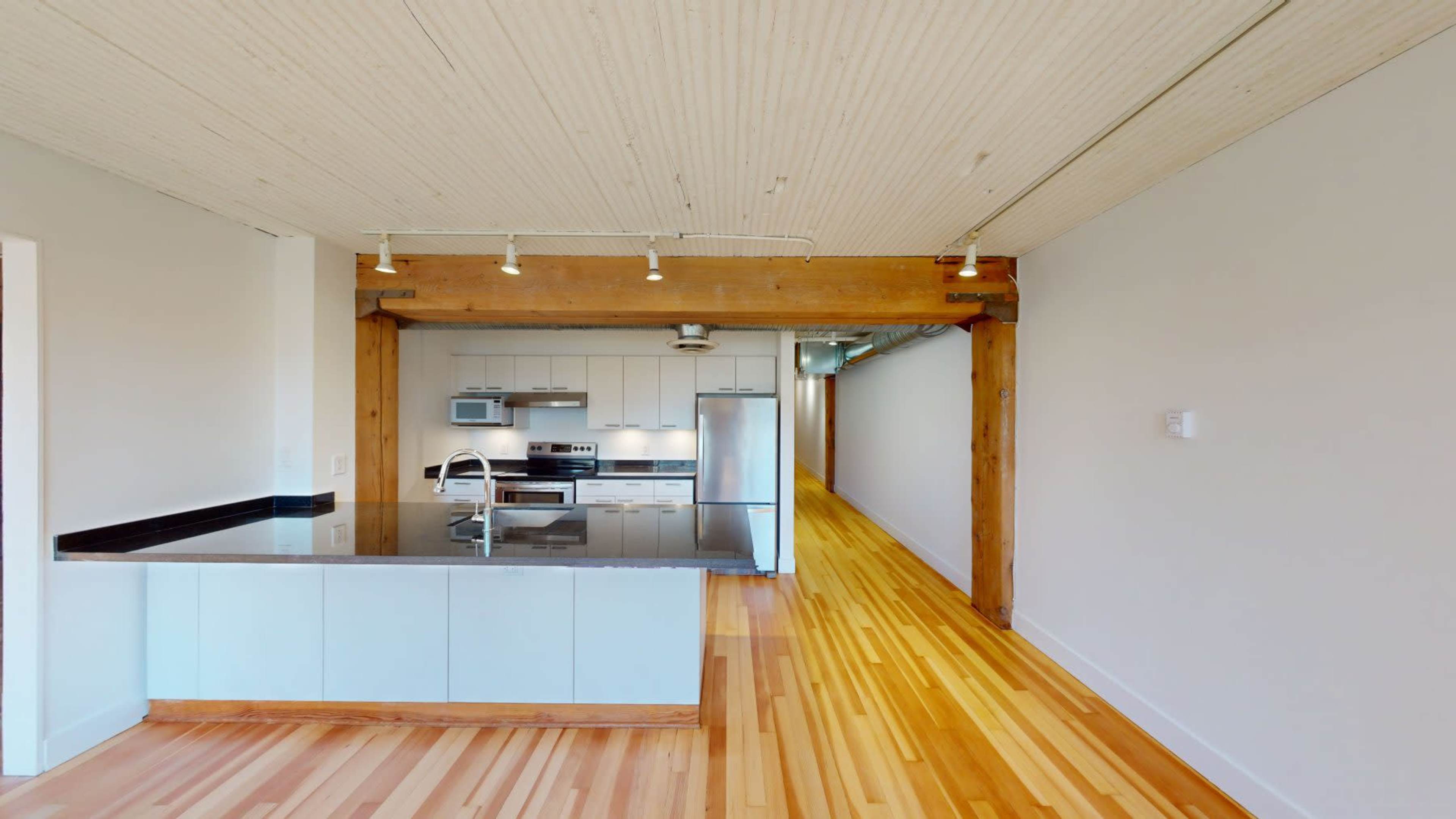 The image shows a modern kitchen with a black countertop, stainless steel appliances, and wooden flooring, connected to an open living space.