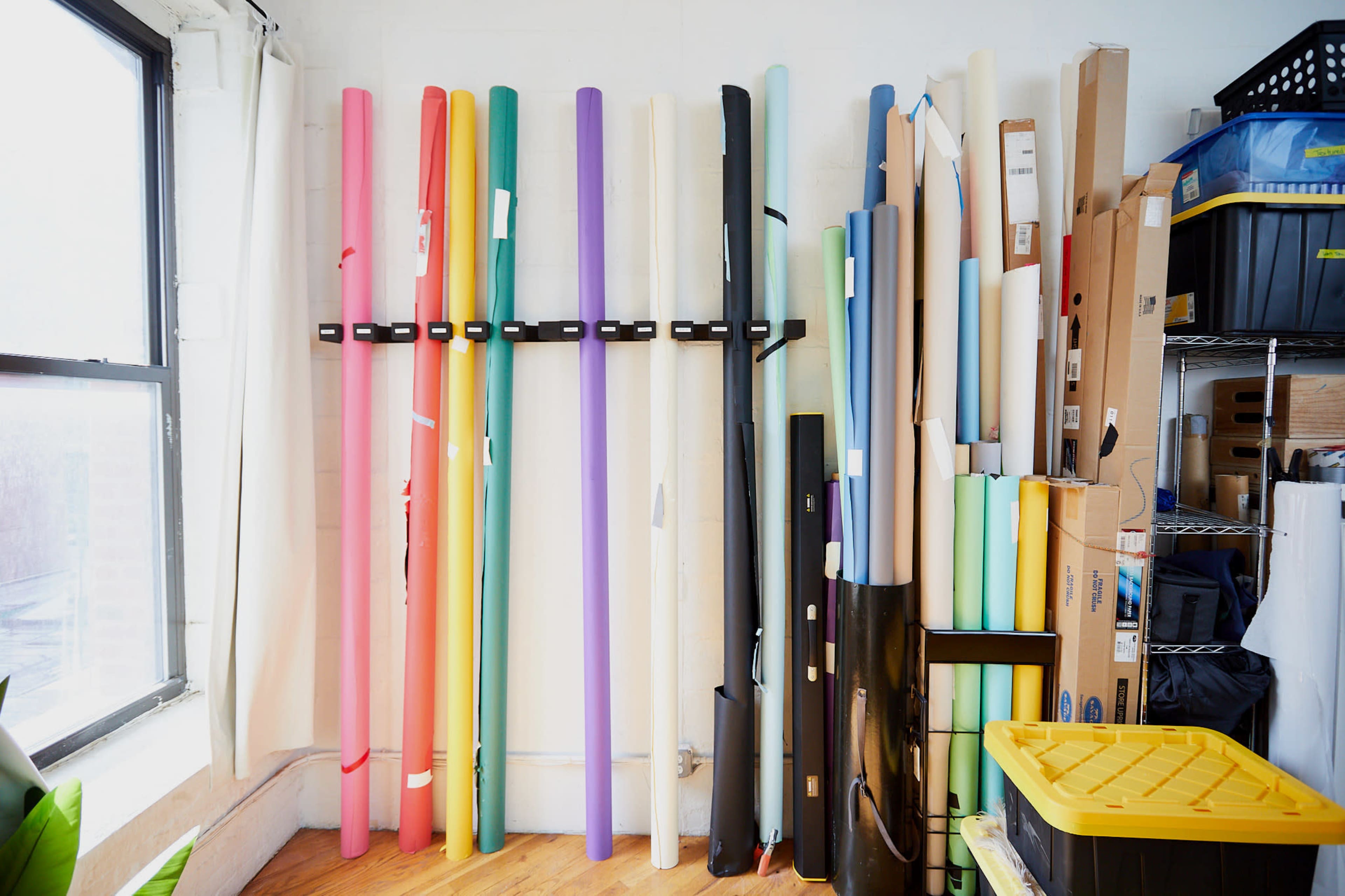 The image shows a collection of rolled-up colored paper and plastic sheets arranged vertically against a wall in a well-lit room.