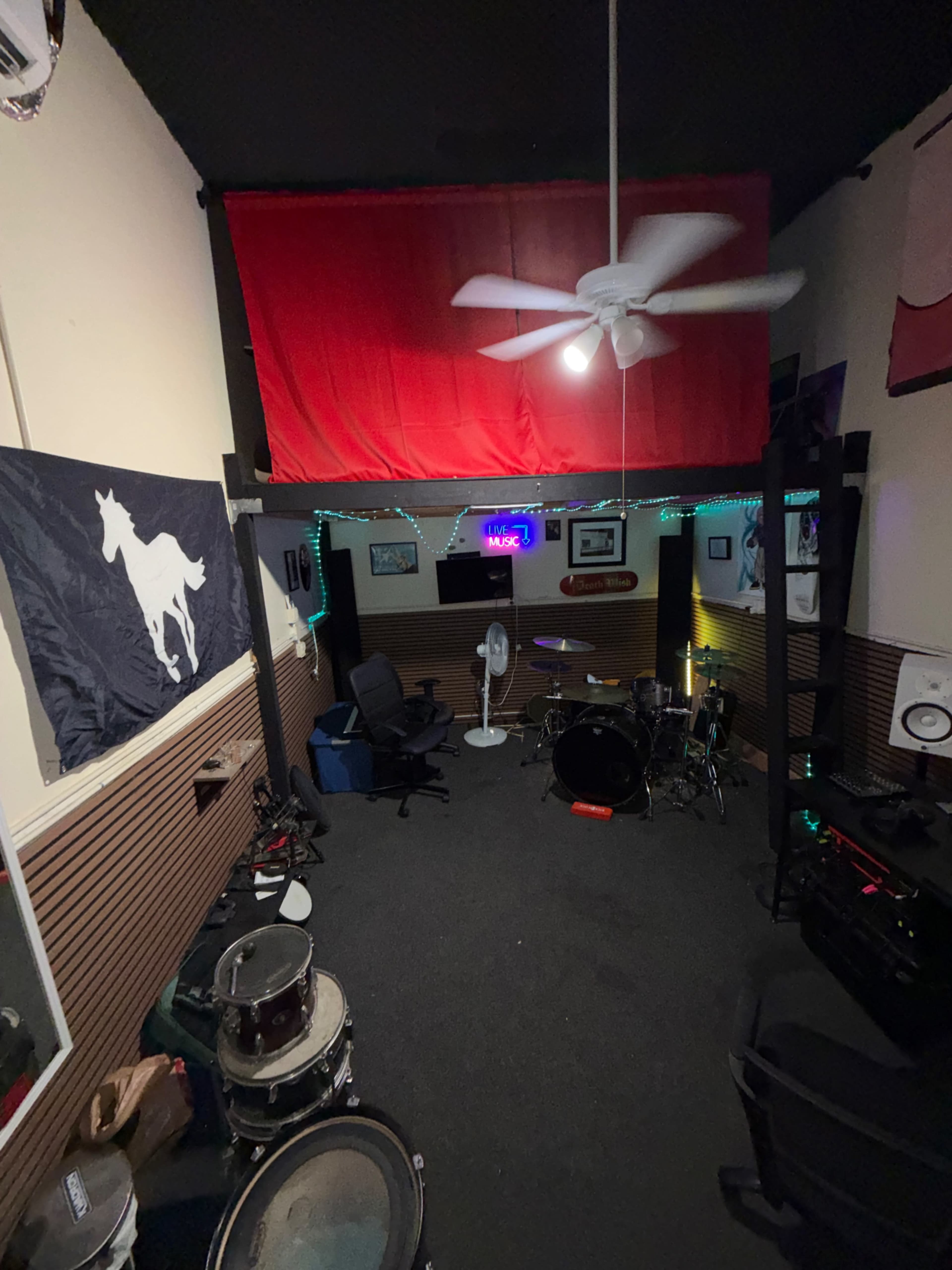 The image shows a music practice room featuring a drum set, a fan, and a red curtain hanging above, with various musical equipment and a horse flag on the wall.