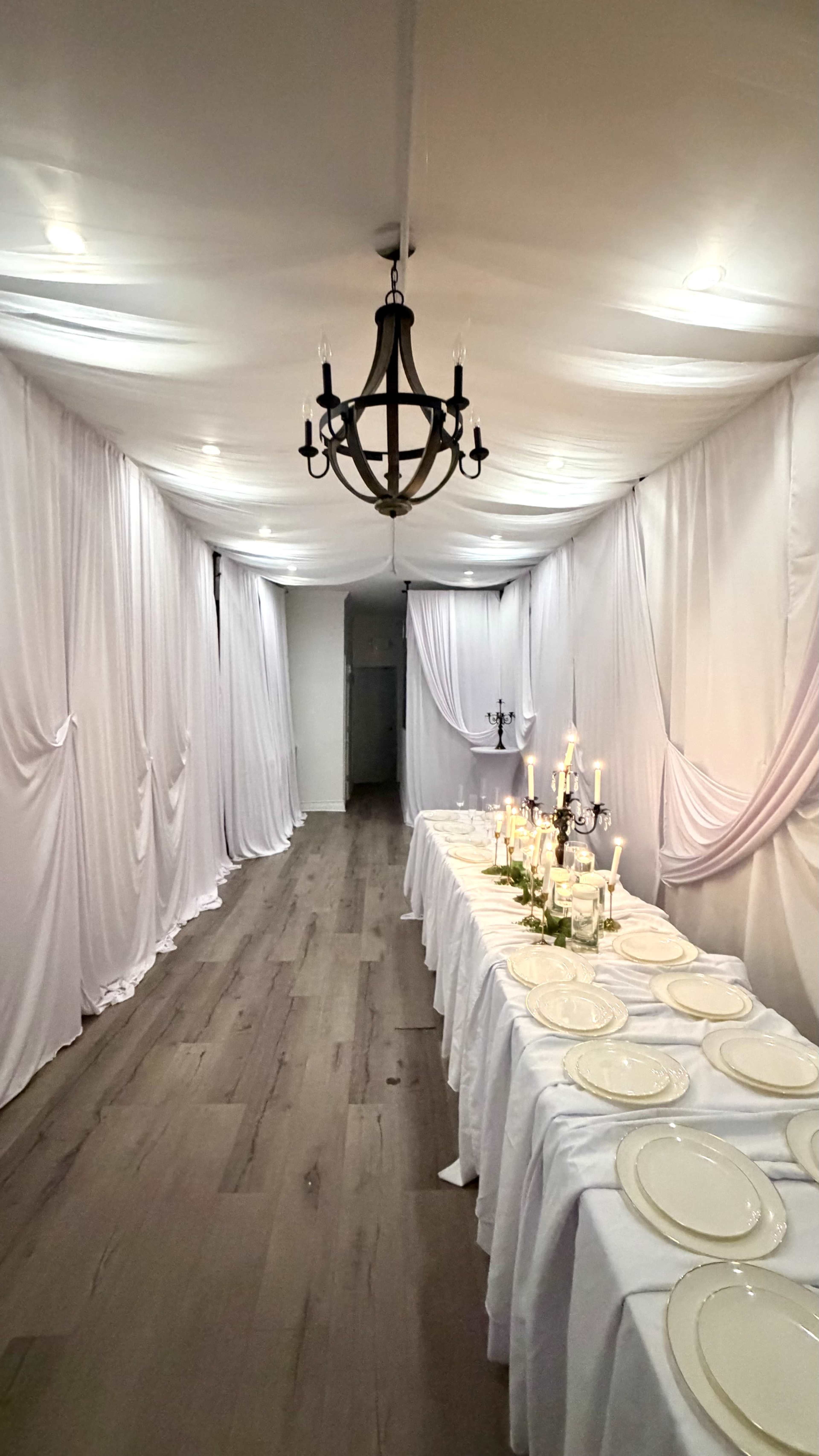 A long, draped hallway features a dining table set with plates and candles under a chandelier.