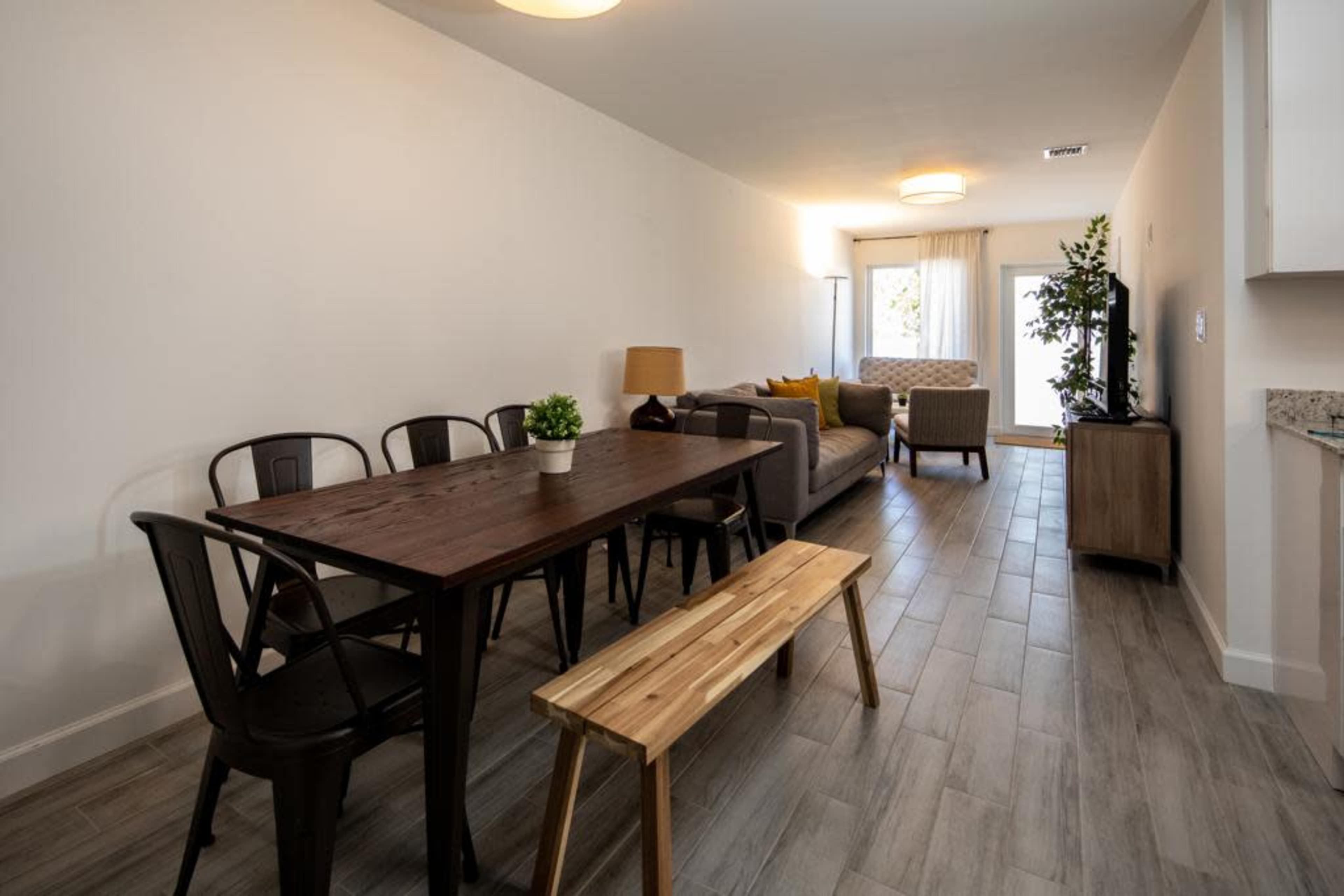 A modern living space featuring a dining table with chairs, a wooden bench, a sofa, and a television, illuminated by natural light.