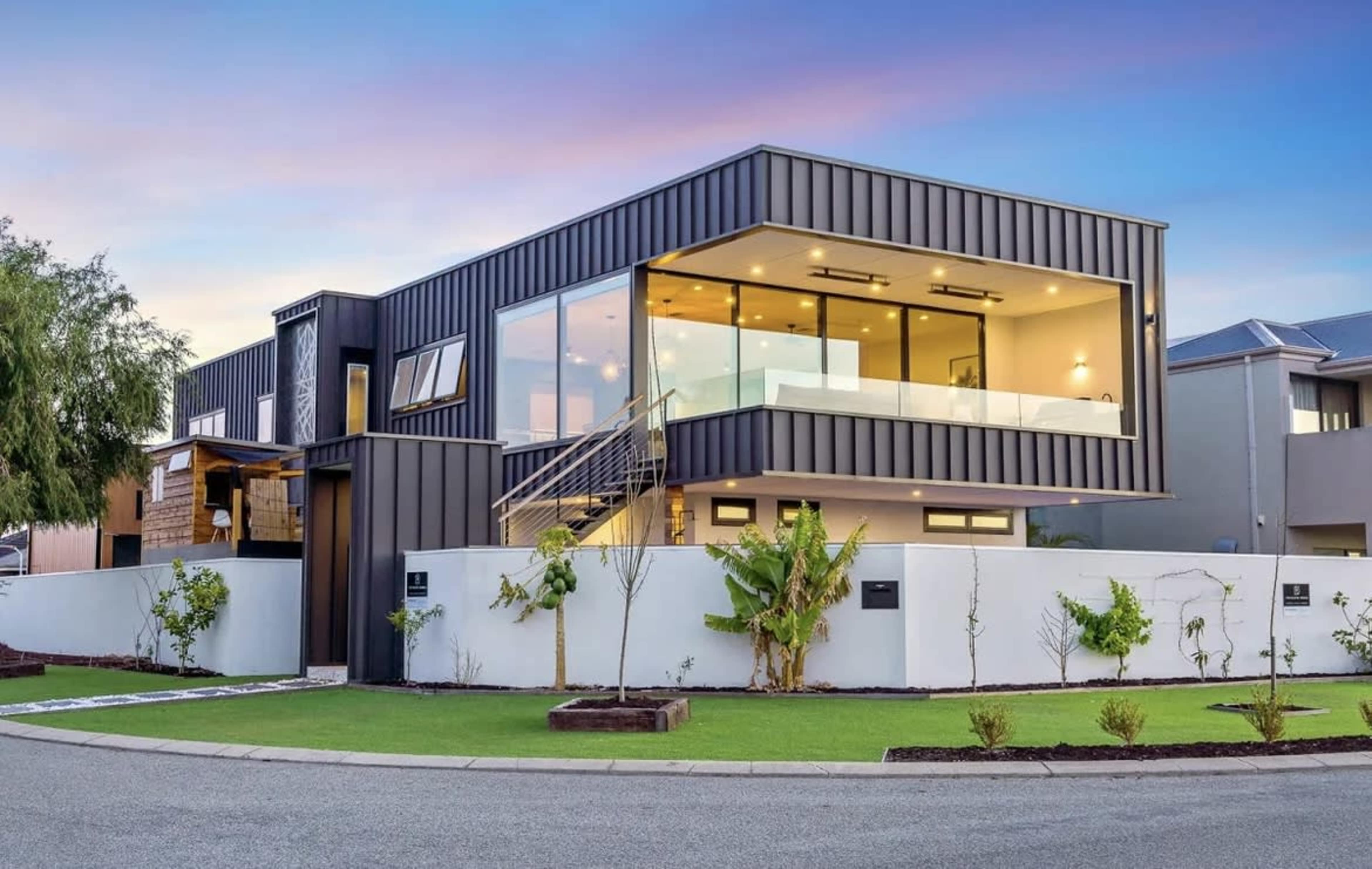 The image shows a modern house with a sleek design and large windows, located at a corner lot with a landscaped yard.