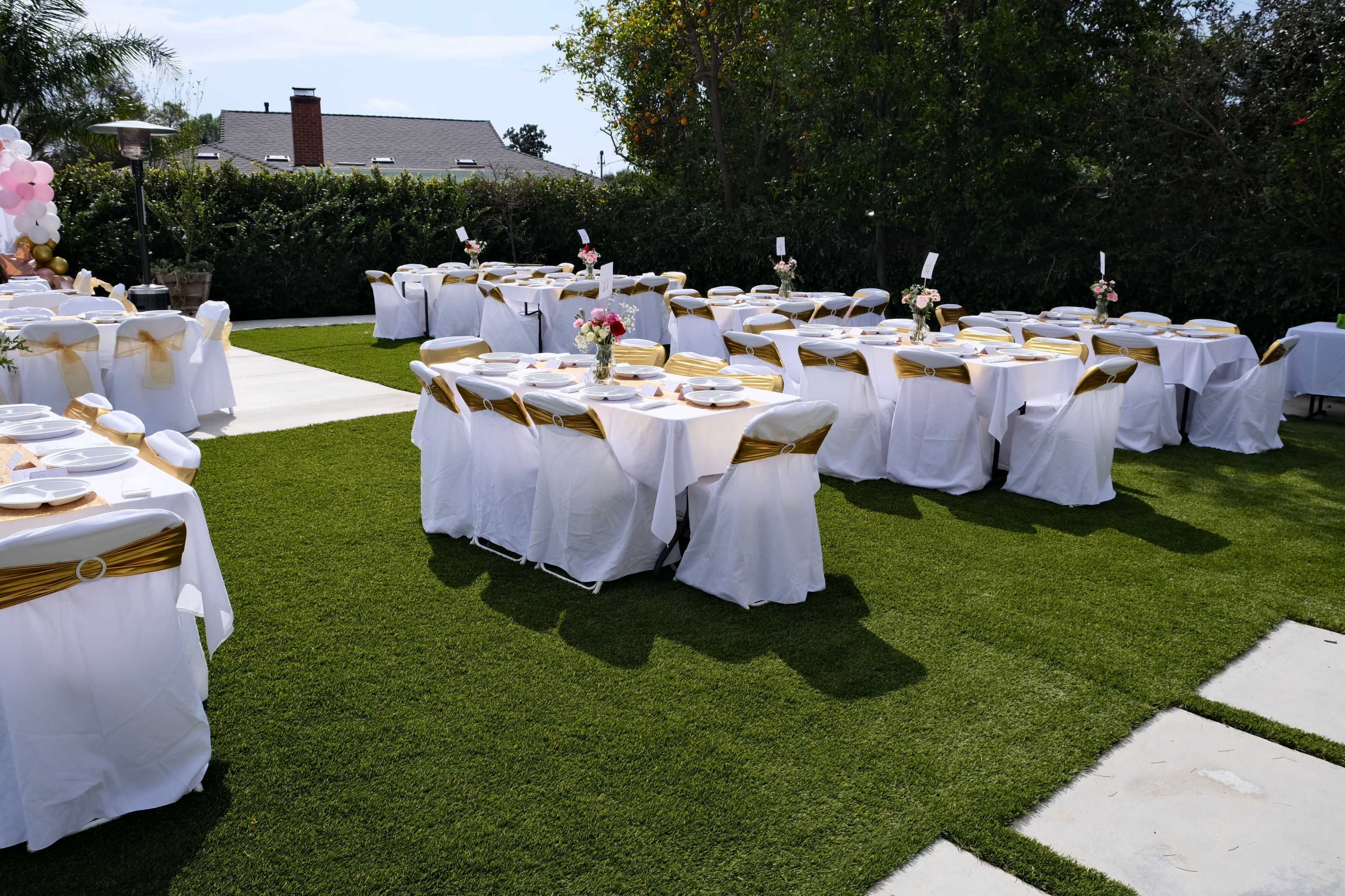 The image shows a garden setting with several tables dressed in white tablecloths and gold accents, arranged for an outdoor event.