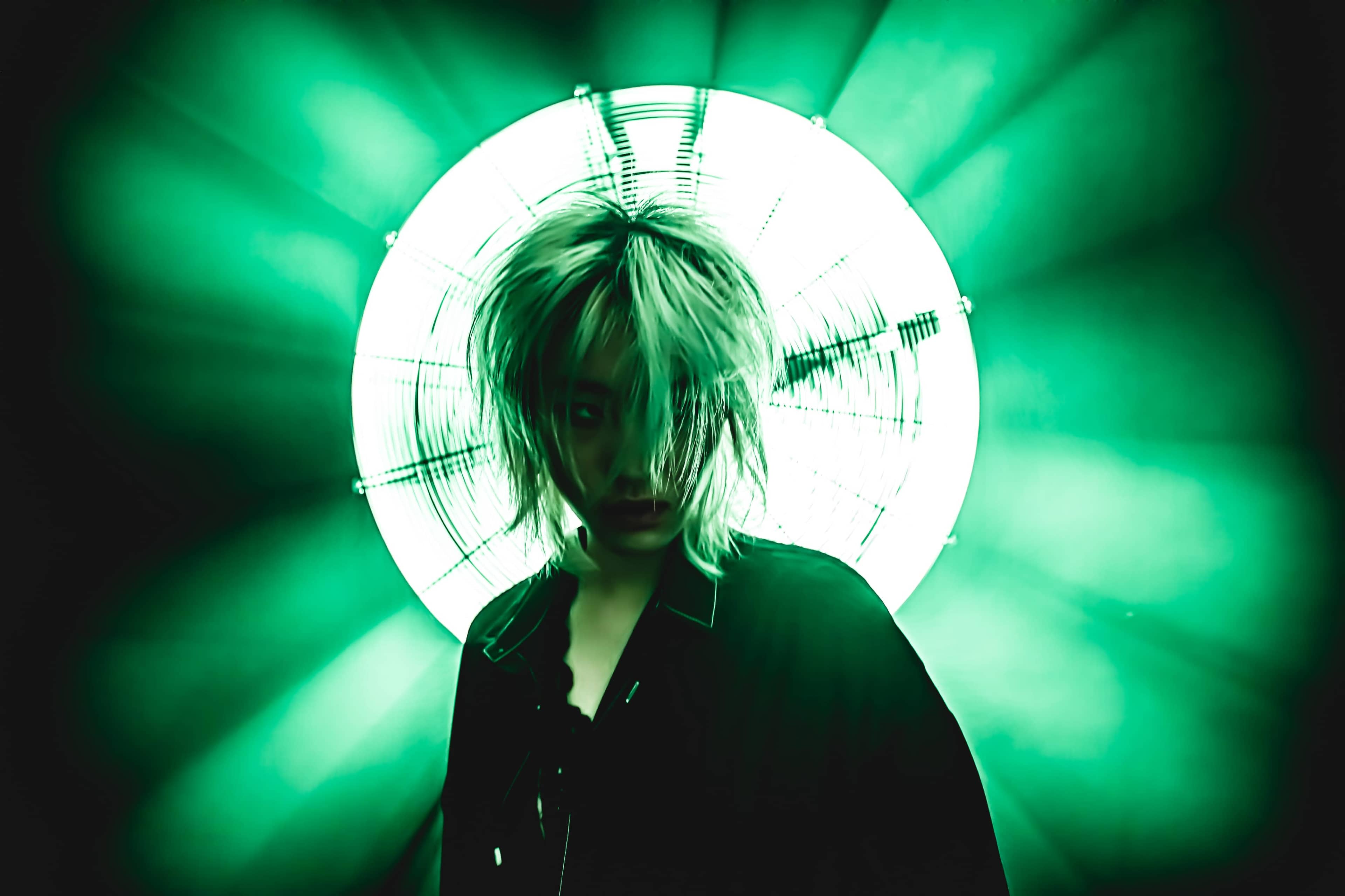 A figure with disheveled hair stands in front of a bright, circular green light source.