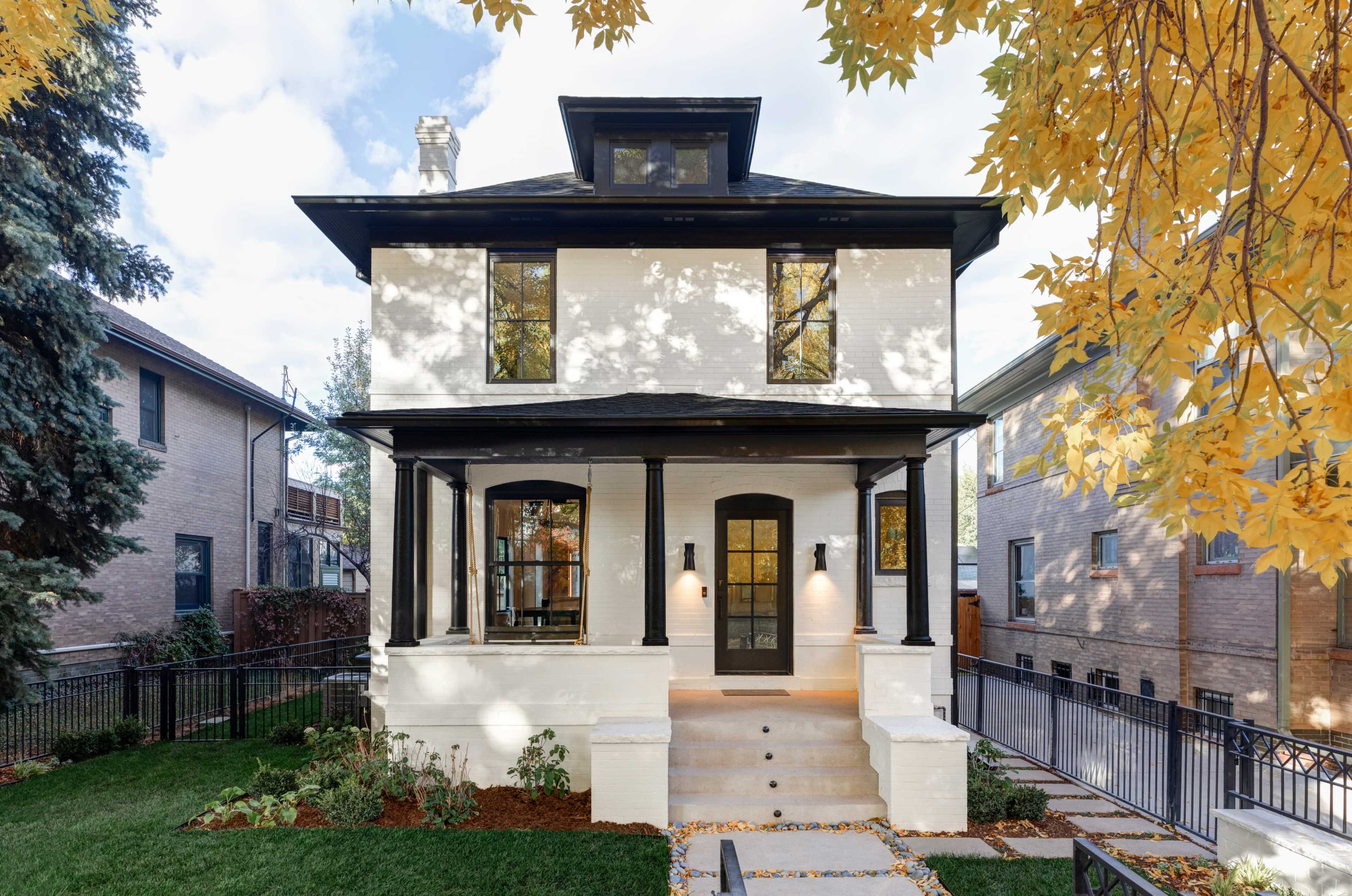 The image shows a two-story, modern-style house with a white exterior, black trim, and a landscaped front yard featuring autumn leaves.