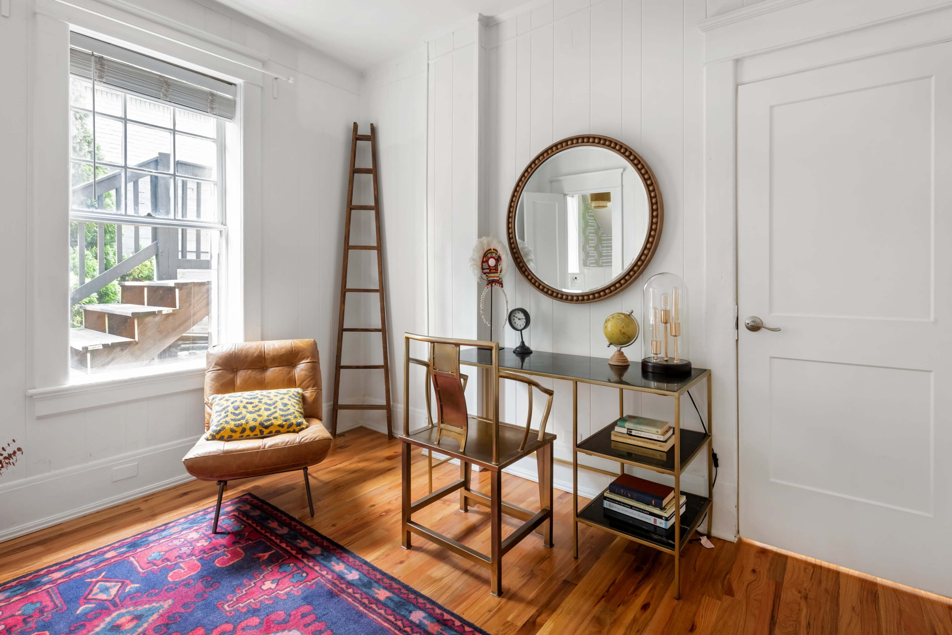 The image shows a modern room with wooden flooring, featuring a styled corner with a chair, a decorative mirror, and a small shelving unit displaying books and a globe.