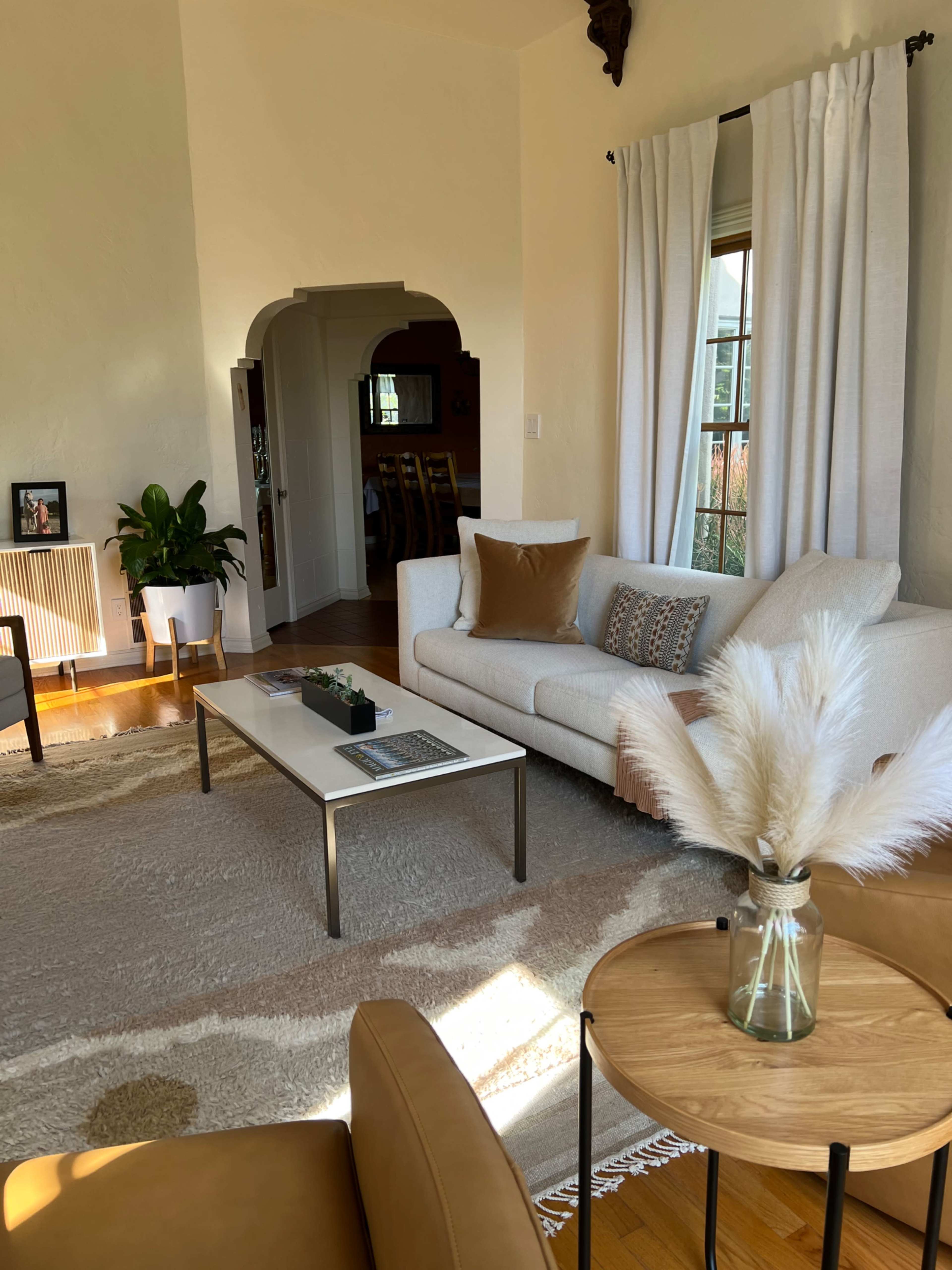 A bright living room features a white sofa, a glass coffee table, and a vase with dried plants, with an archway leading to a dining area in the background.
