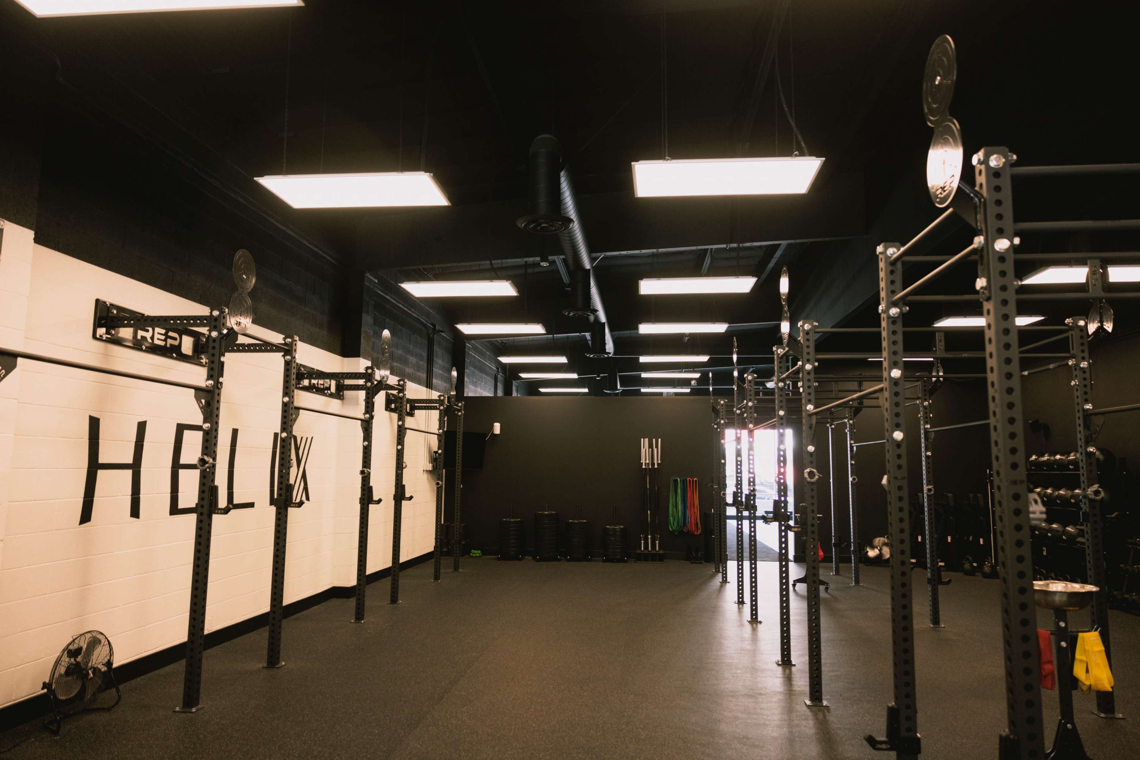 The image shows a spacious, well-lit gym with multiple weightlifting racks and exercise equipment against black walls.