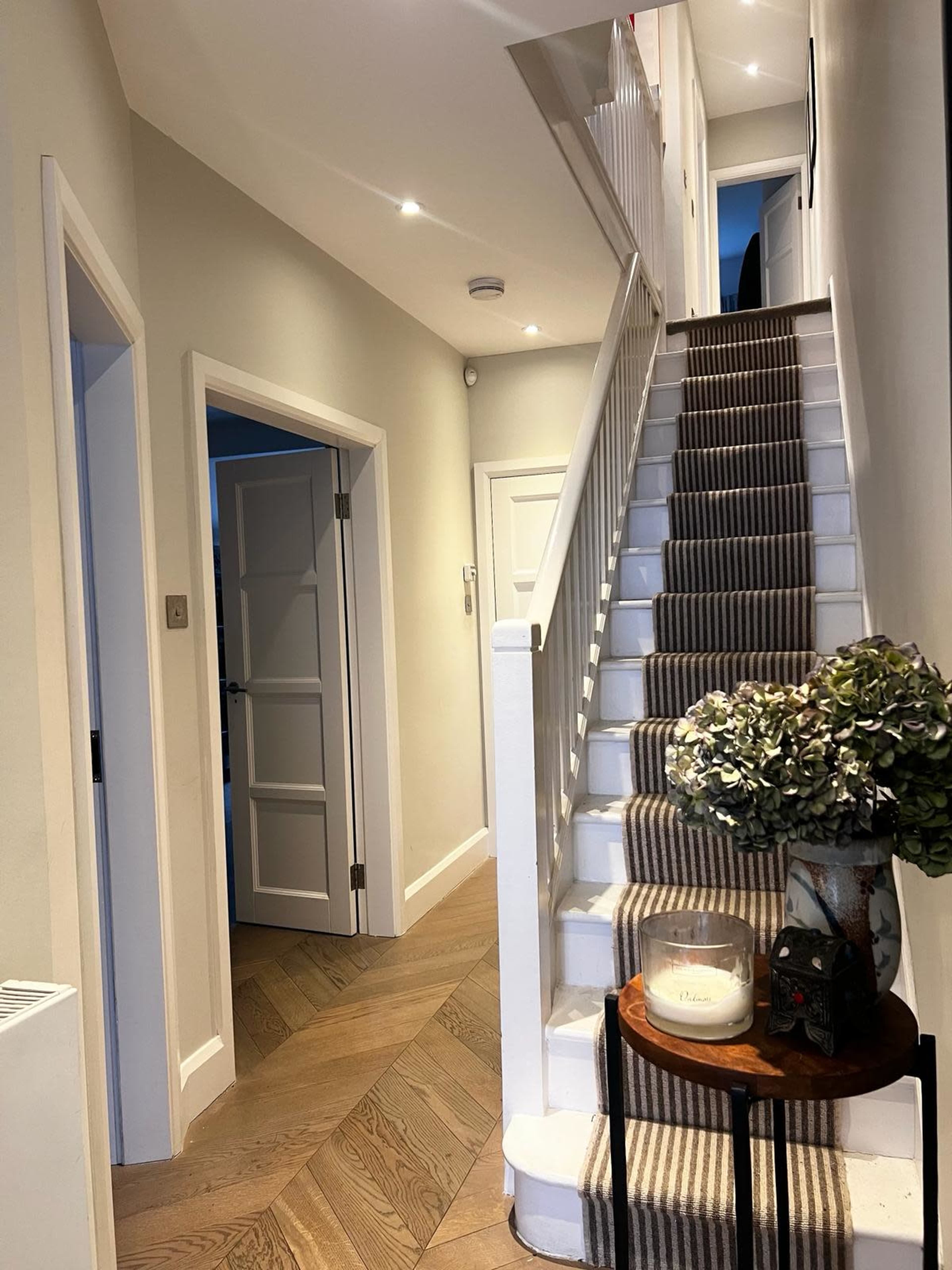 The image shows a narrow hallway with a staircase featuring striped carpet, leading to an upper level, and doorways on the left.
