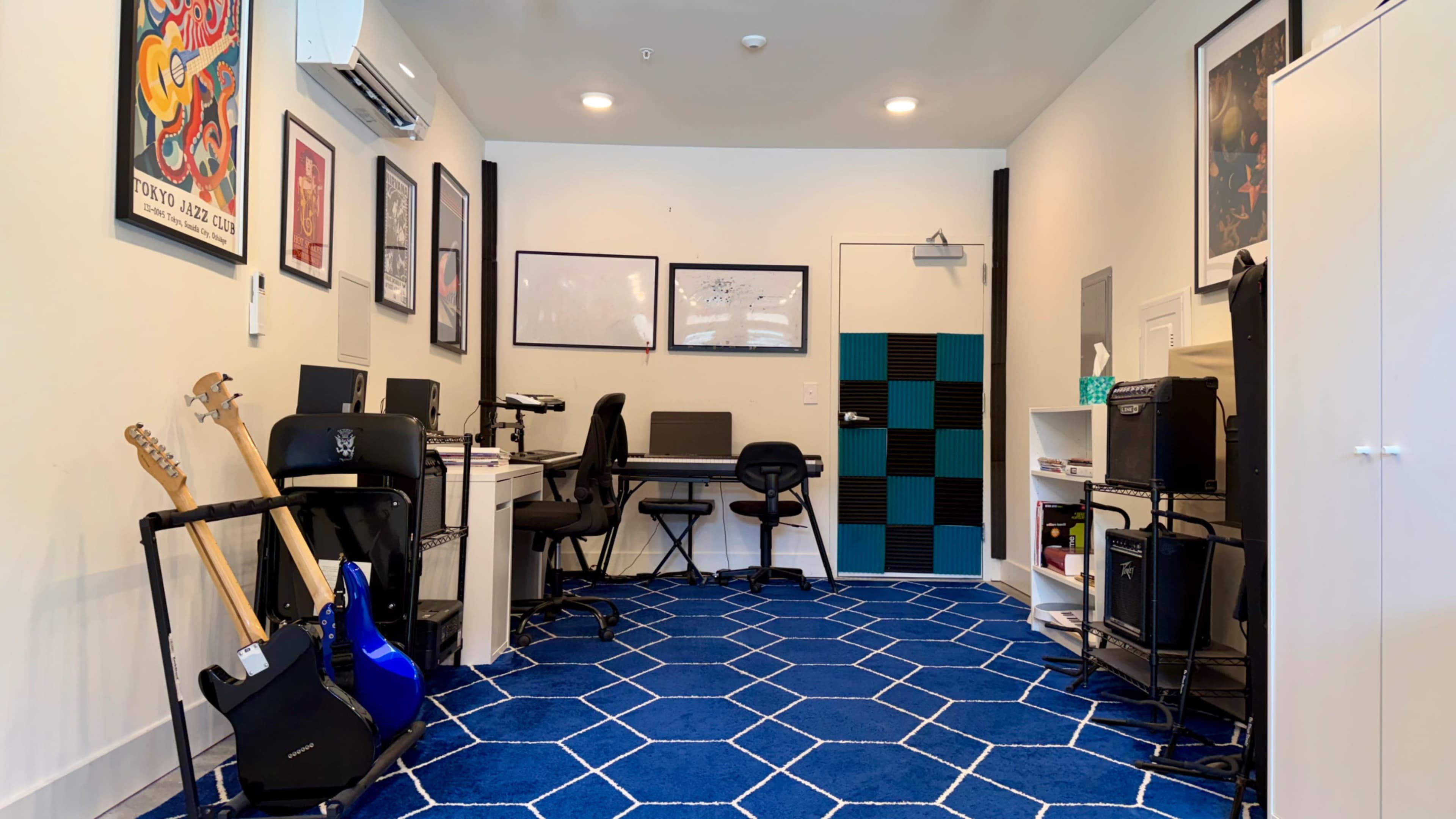 The image shows a modern office space with a blue geometric carpet, two desks with computers, musical instruments, and framed artwork on the walls.