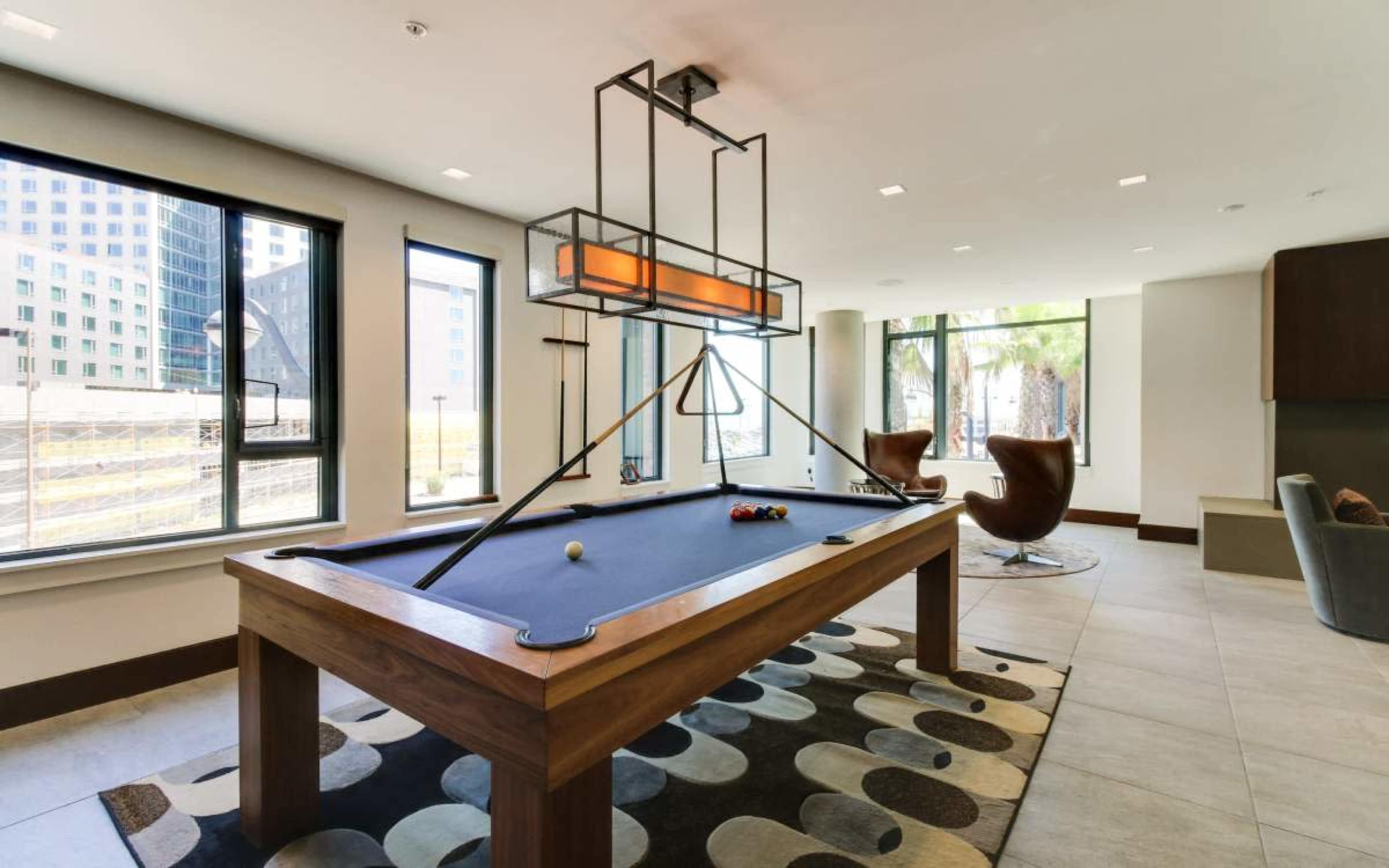 The image shows a modern game room featuring a pool table, stylish lighting, and large windows overlooking an outdoor area.