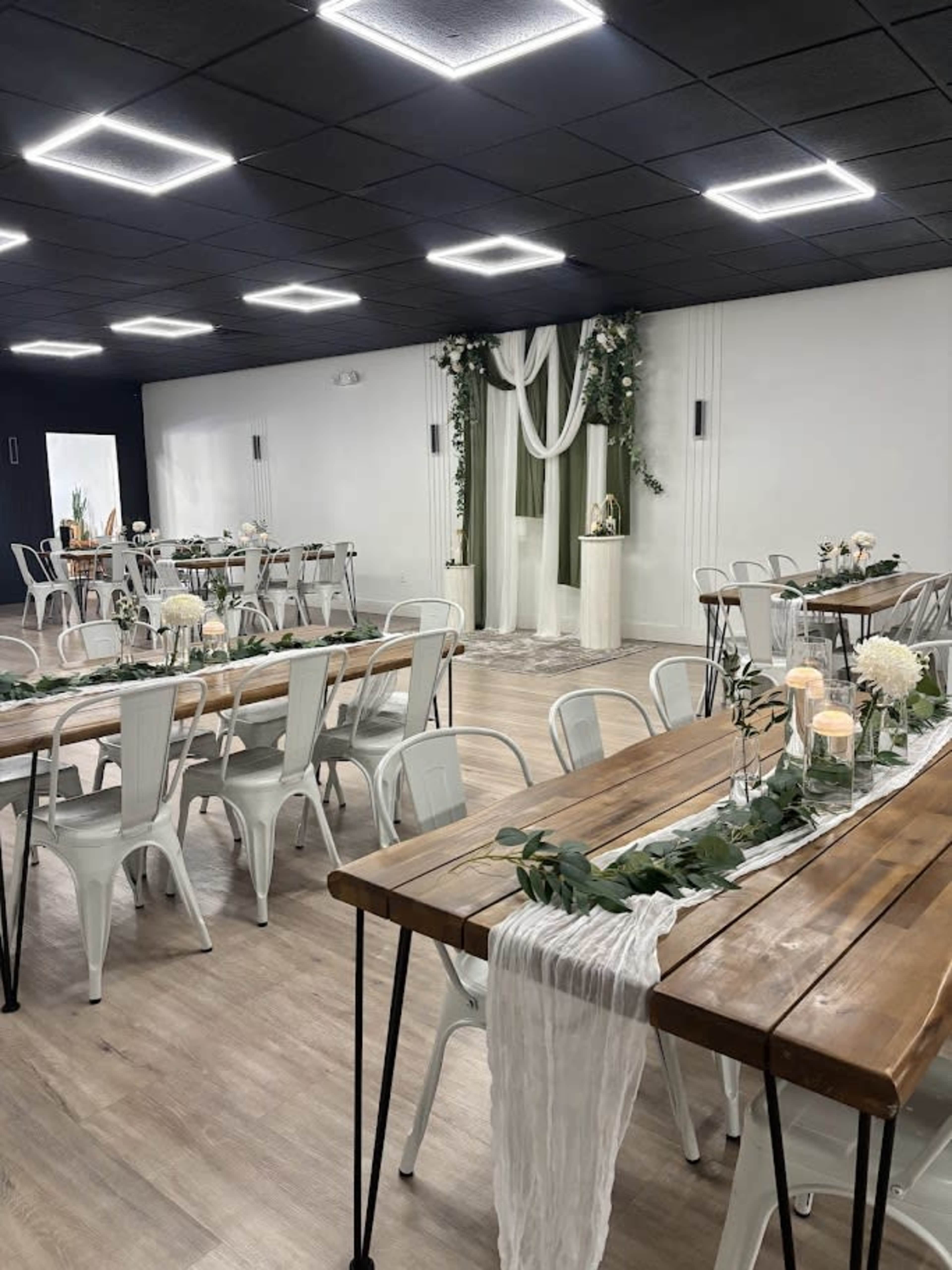 The image shows an event space arranged with wooden tables and white chairs, decorated with greenery and floral centerpieces.