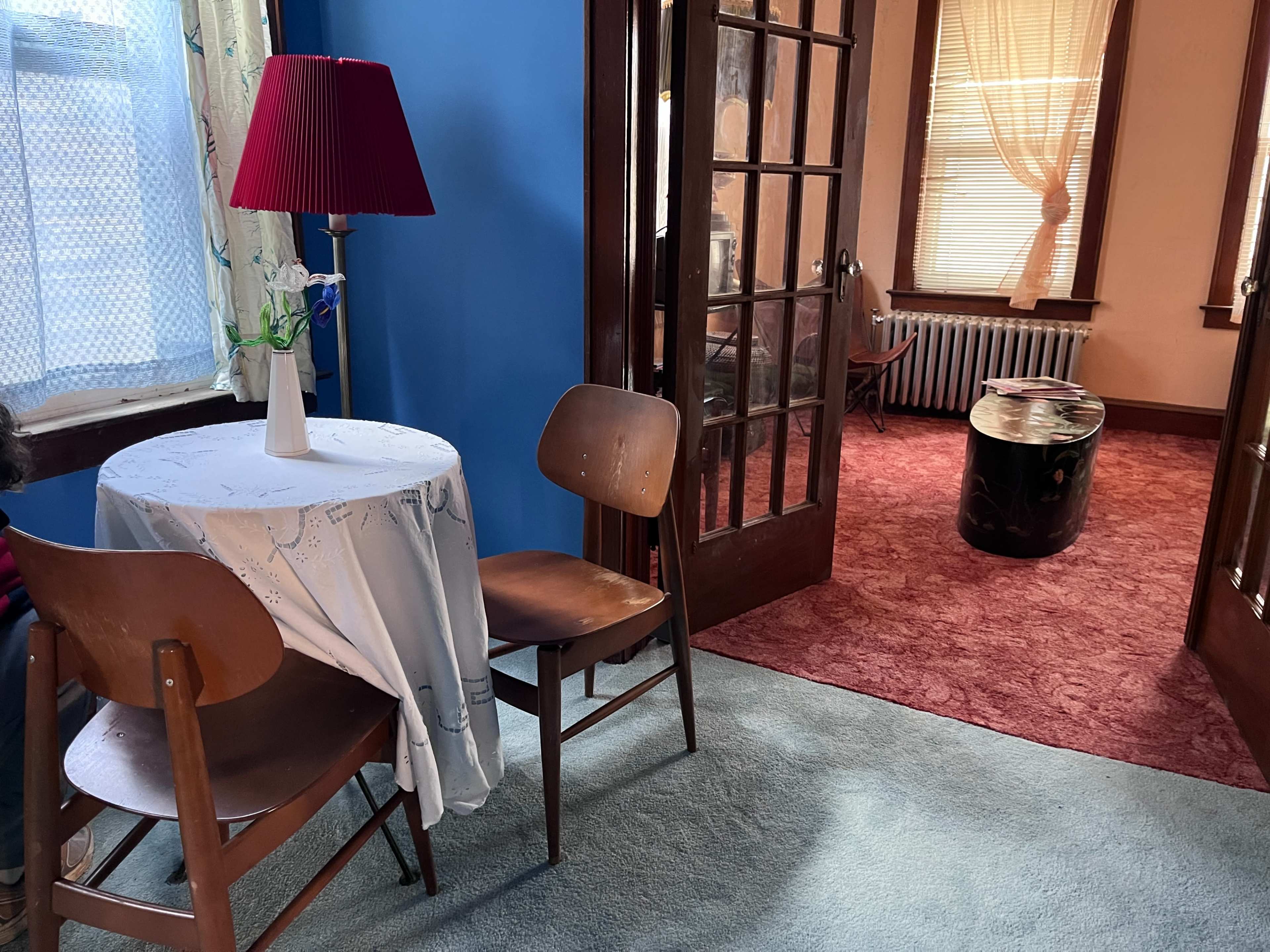 The image shows a small room with a table and two chairs, a red lamp on the table, and a doorway leading to another room with a patterned carpet.