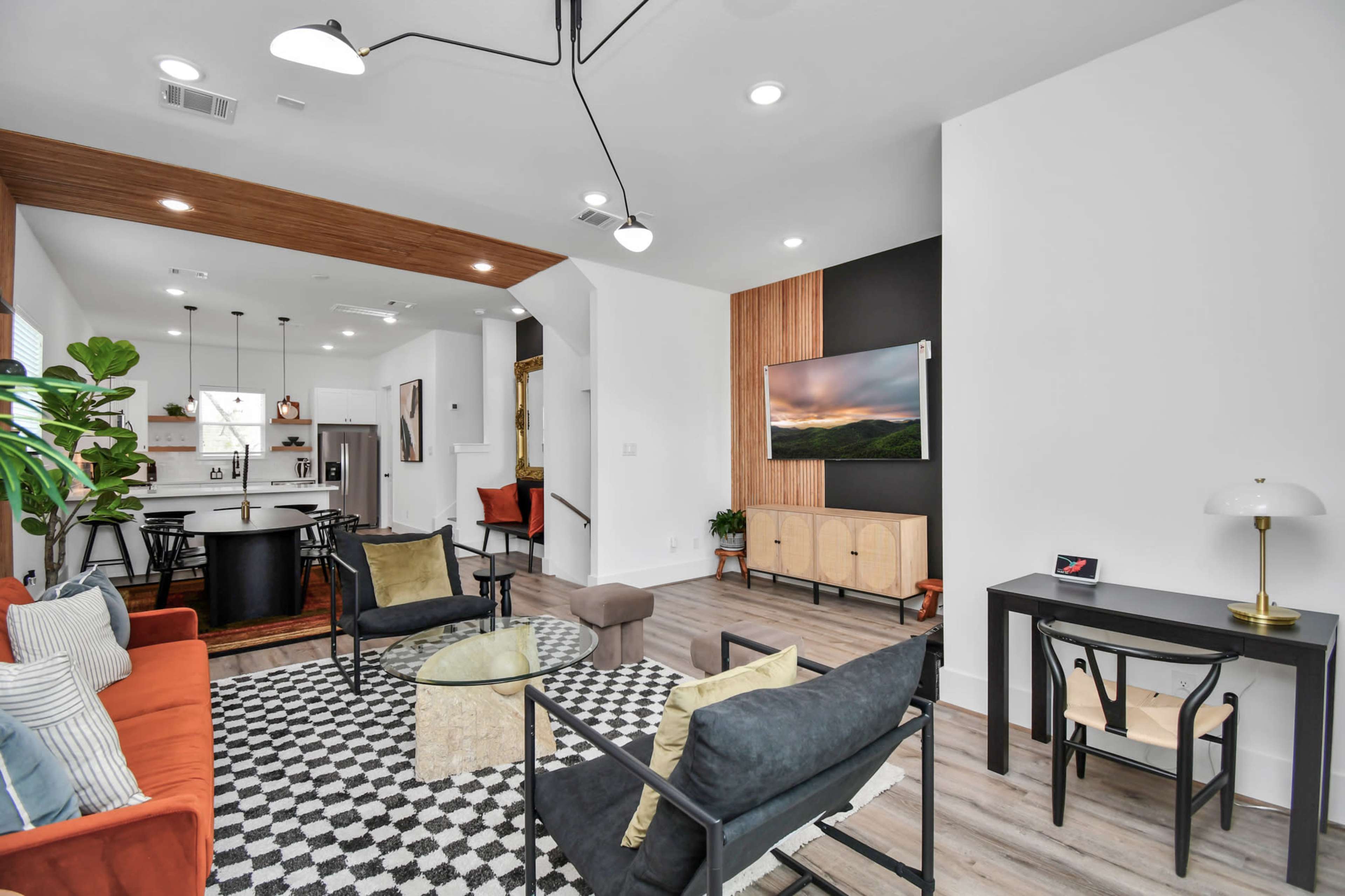 The image shows a modern living room featuring a mix of seating options, a television mounted on the wall, and a dining area visible in the background.