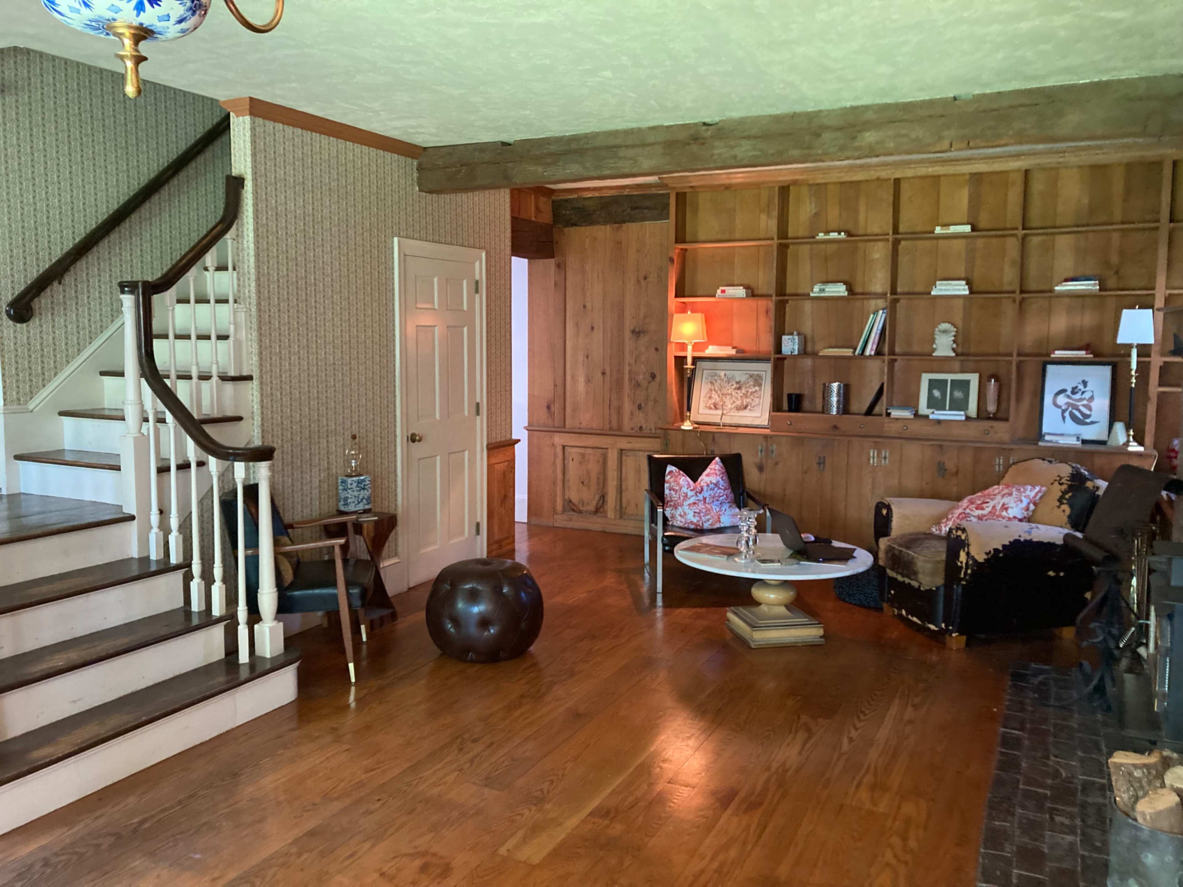 A spacious living room with a staircase on the left, a wooden shelving unit against the wall, and a circular coffee table in the center surrounded by a couch and seating.