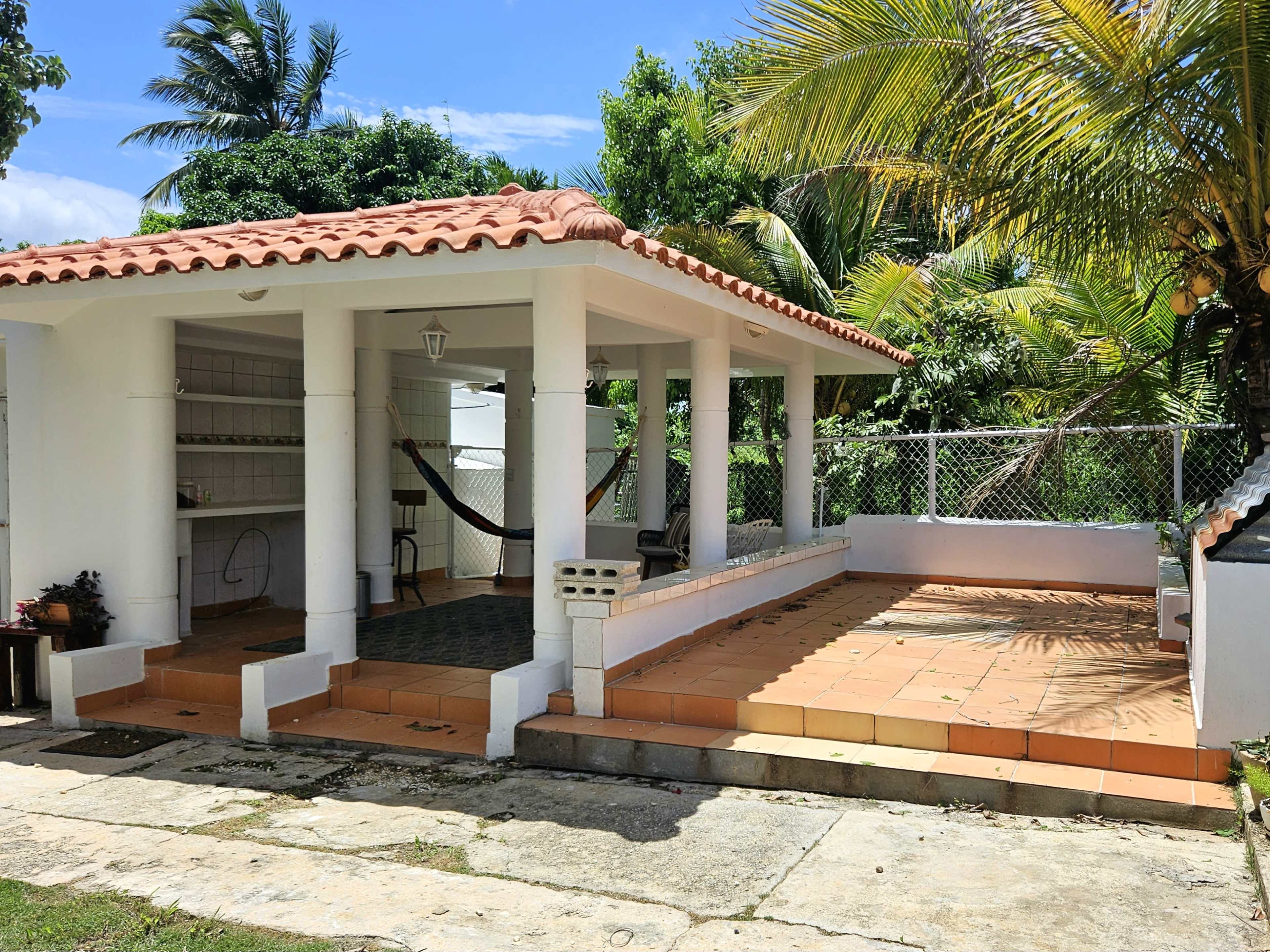 The image shows a tropical outdoor patio area with a tiled floor, a hammock, and a house with a red-tiled roof surrounded by lush greenery.