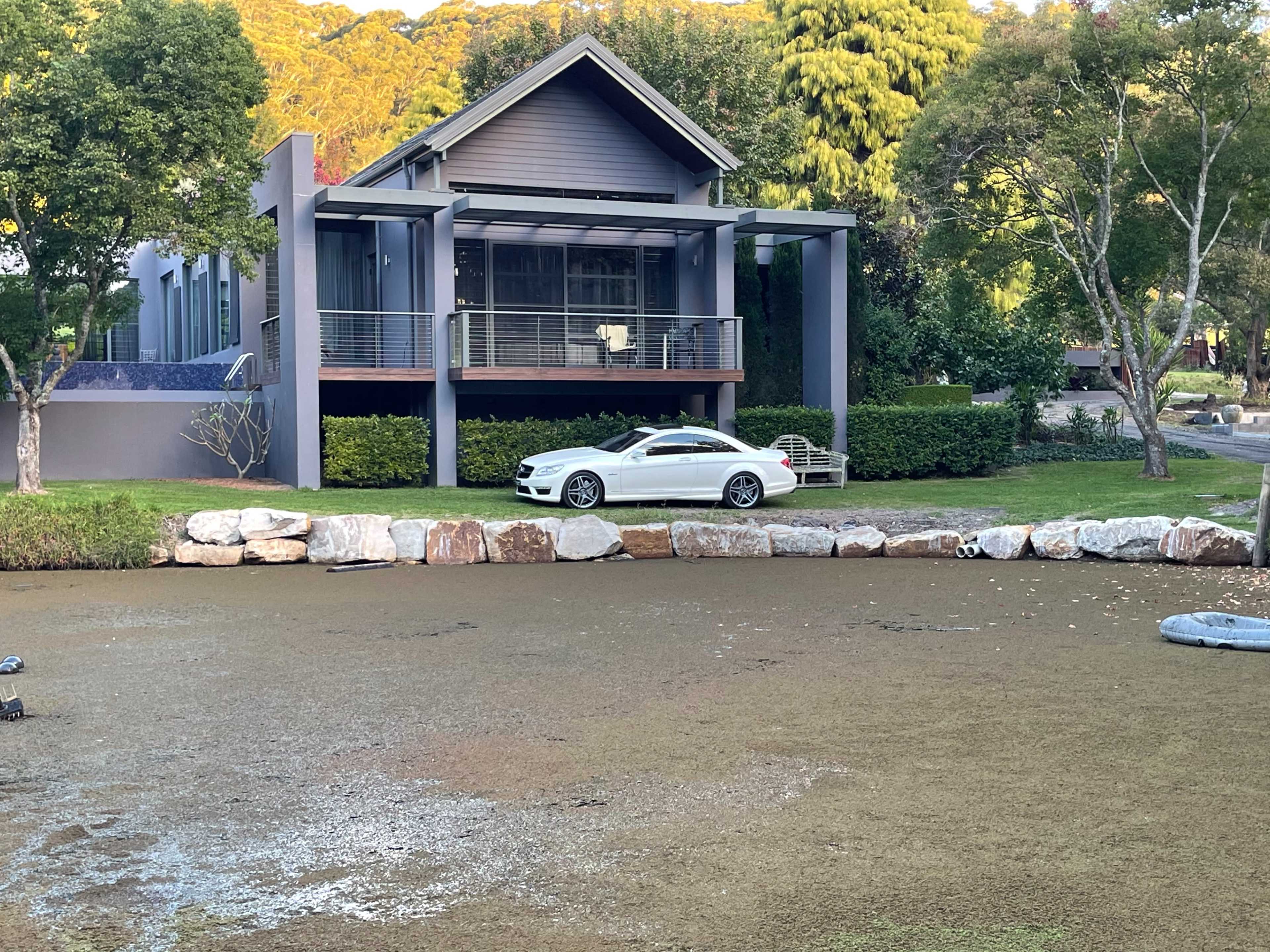 A modern house with a car parked in front is situated by a waterway lined with rocks and greenery.