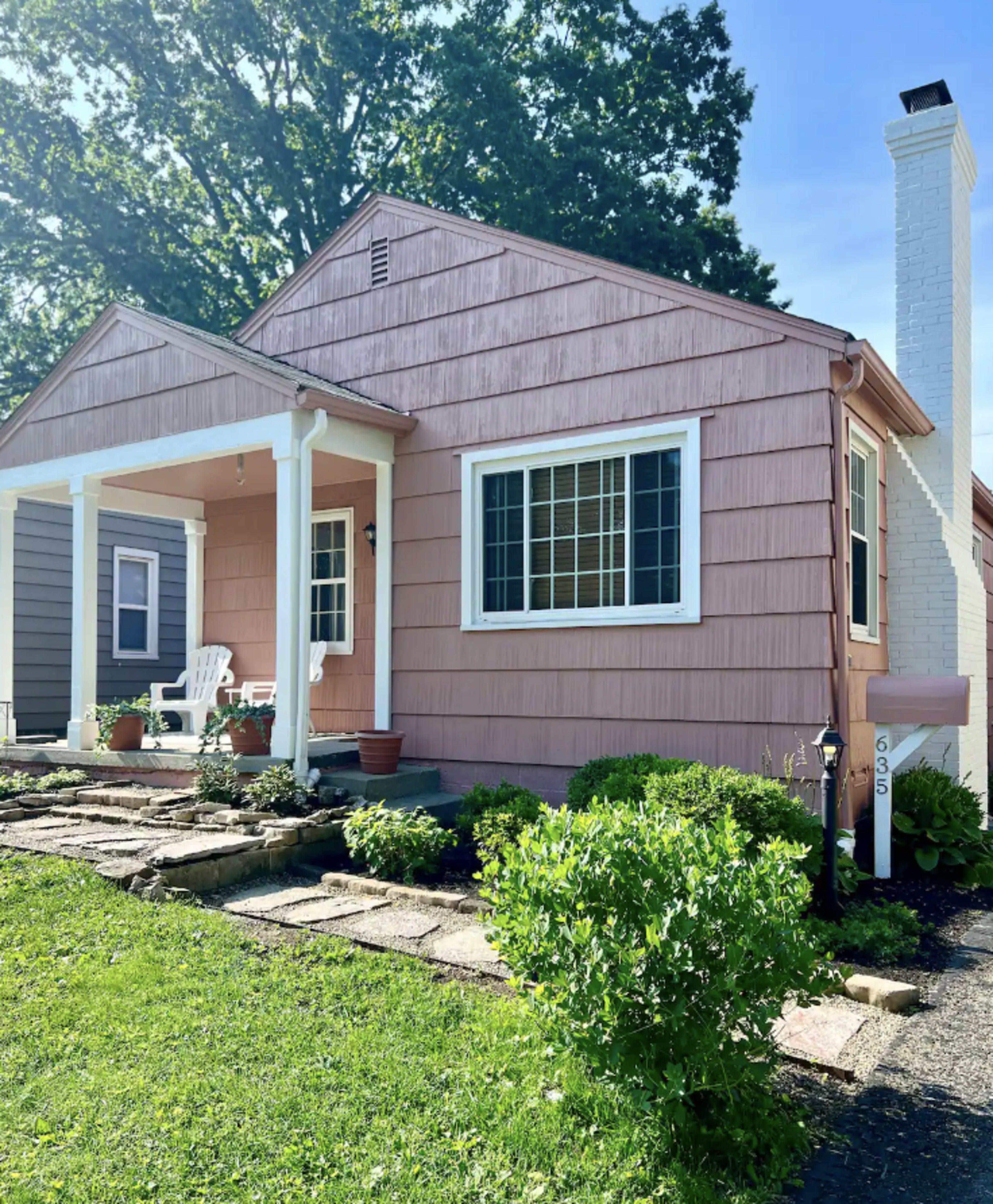 A pink, single-story house features a front porch with white chairs and is surrounded by a landscaped yard.