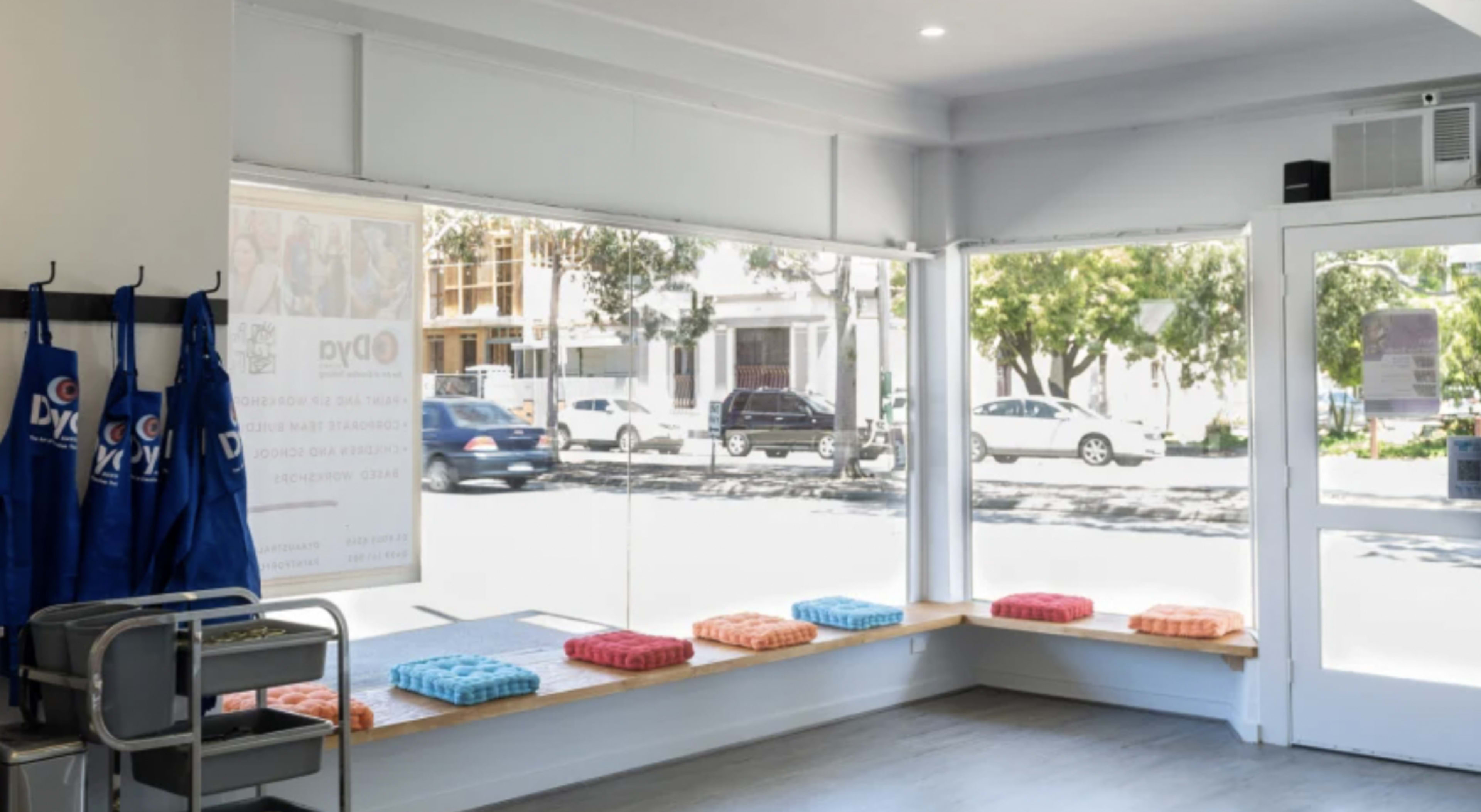 The image shows a bright, modern waiting area with large windows, benches lined with colorful cushions, and a cart in the corner.