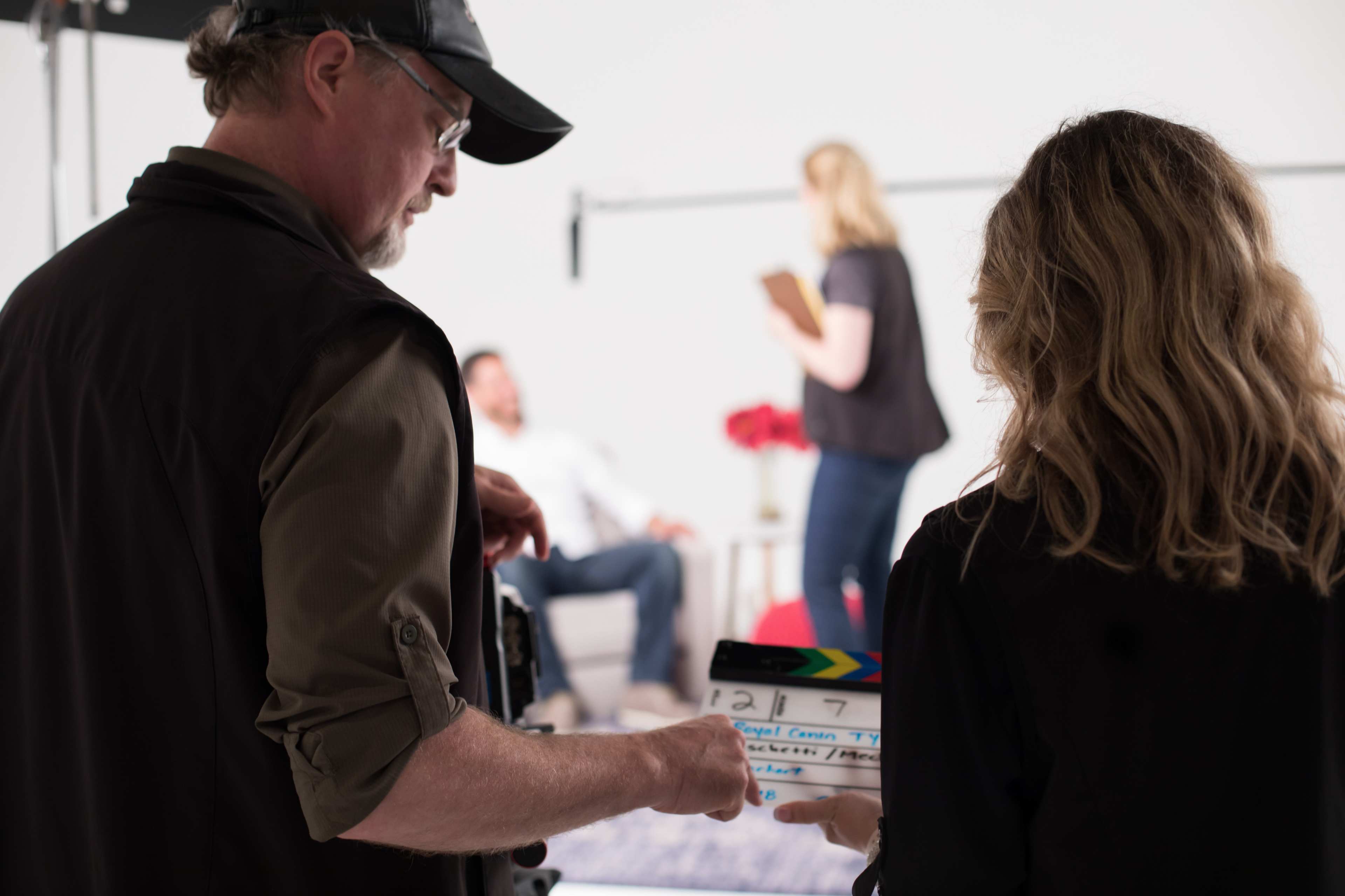 A man and a woman prepare to film a scene with a clapboard in a studio setting, while two actors are visible in the background.