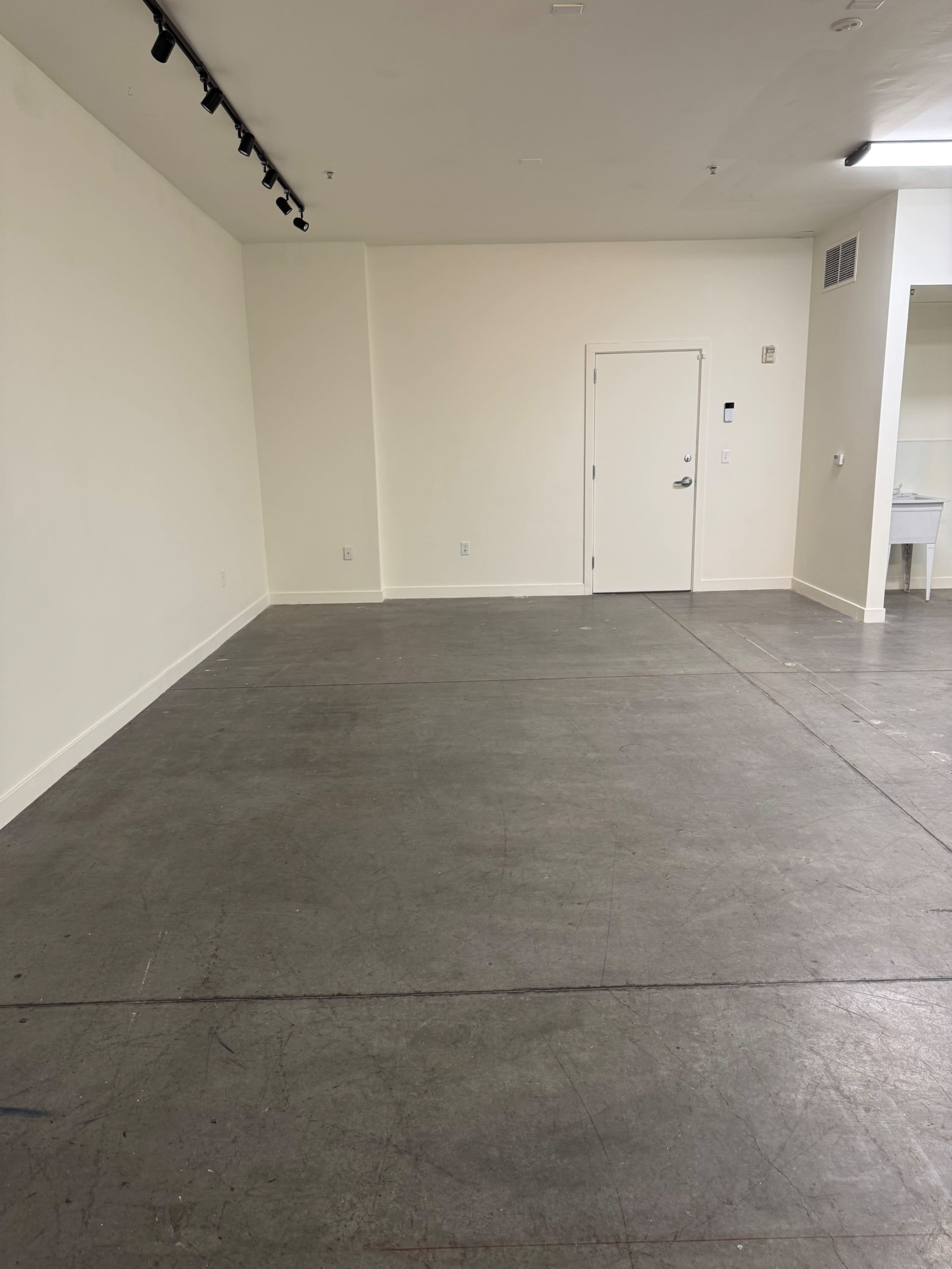 The image shows an empty room with a concrete floor, white walls, and a closed door.