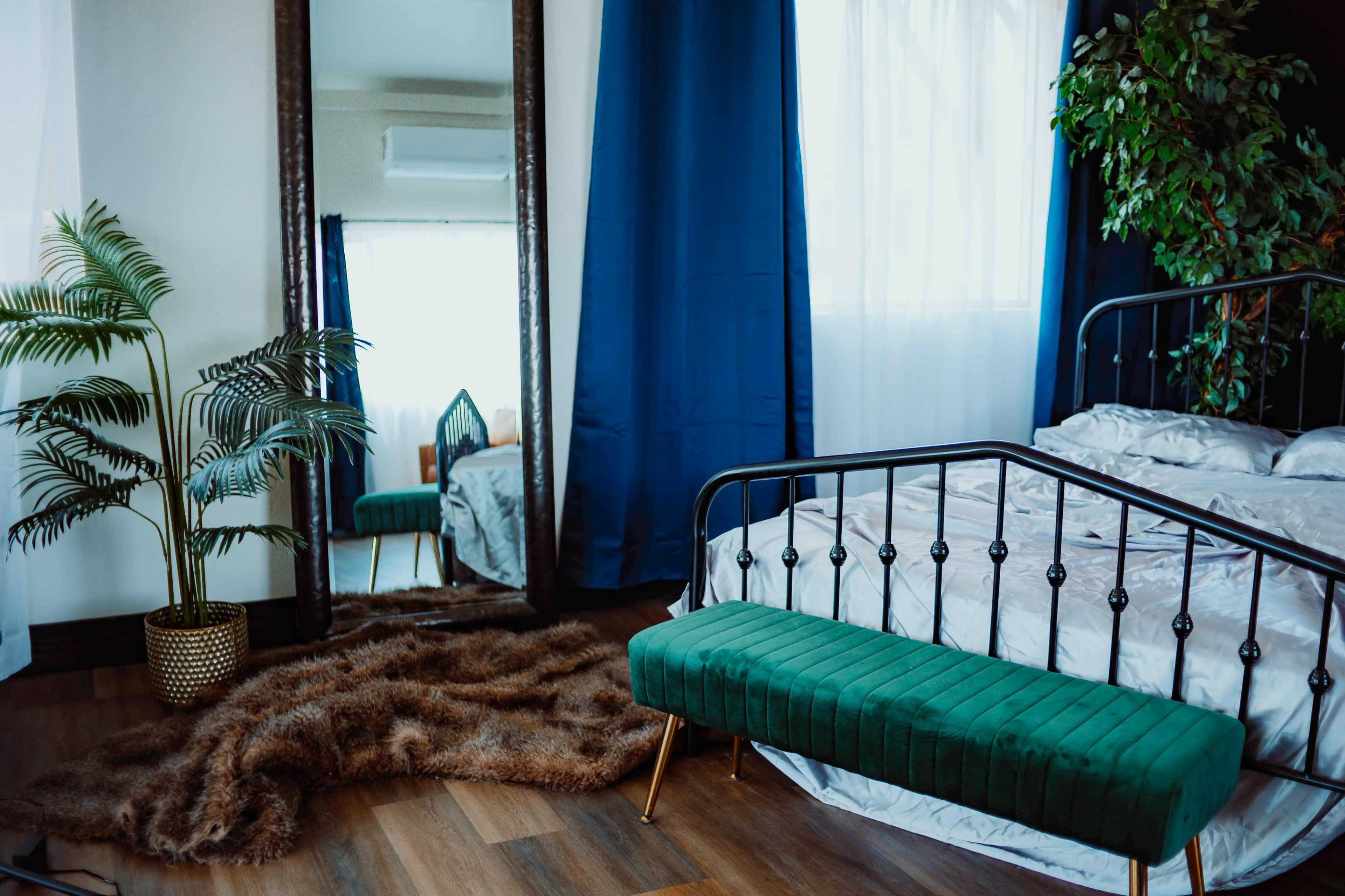 A modern bedroom features a green bench at the foot of a bed, a large mirror, a potted plant, and a textured rug on a wooden floor.