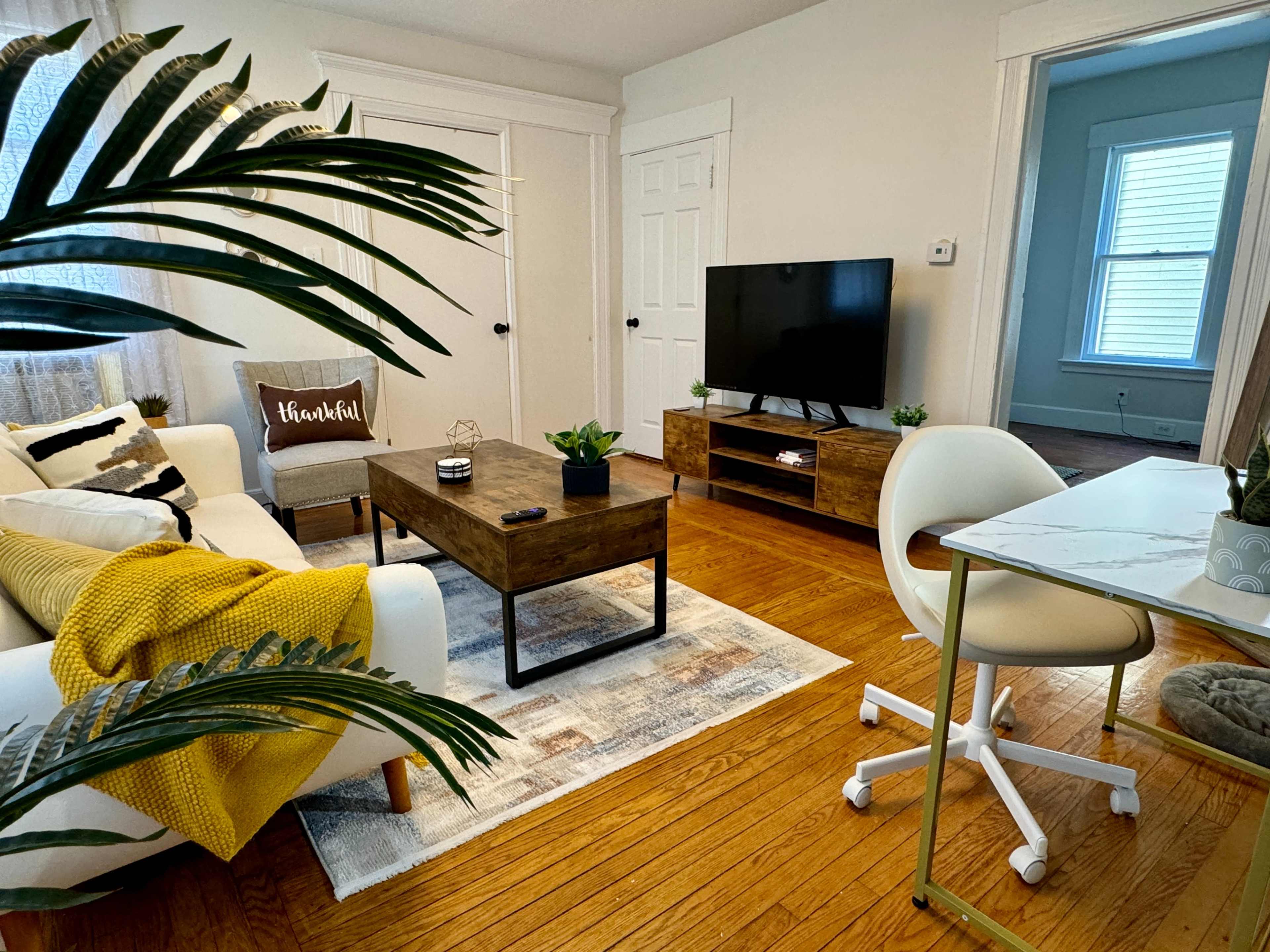 A cozy living room with a white couch, a wooden coffee table, a television on a wooden stand, and a small workspace with a desk and chair.