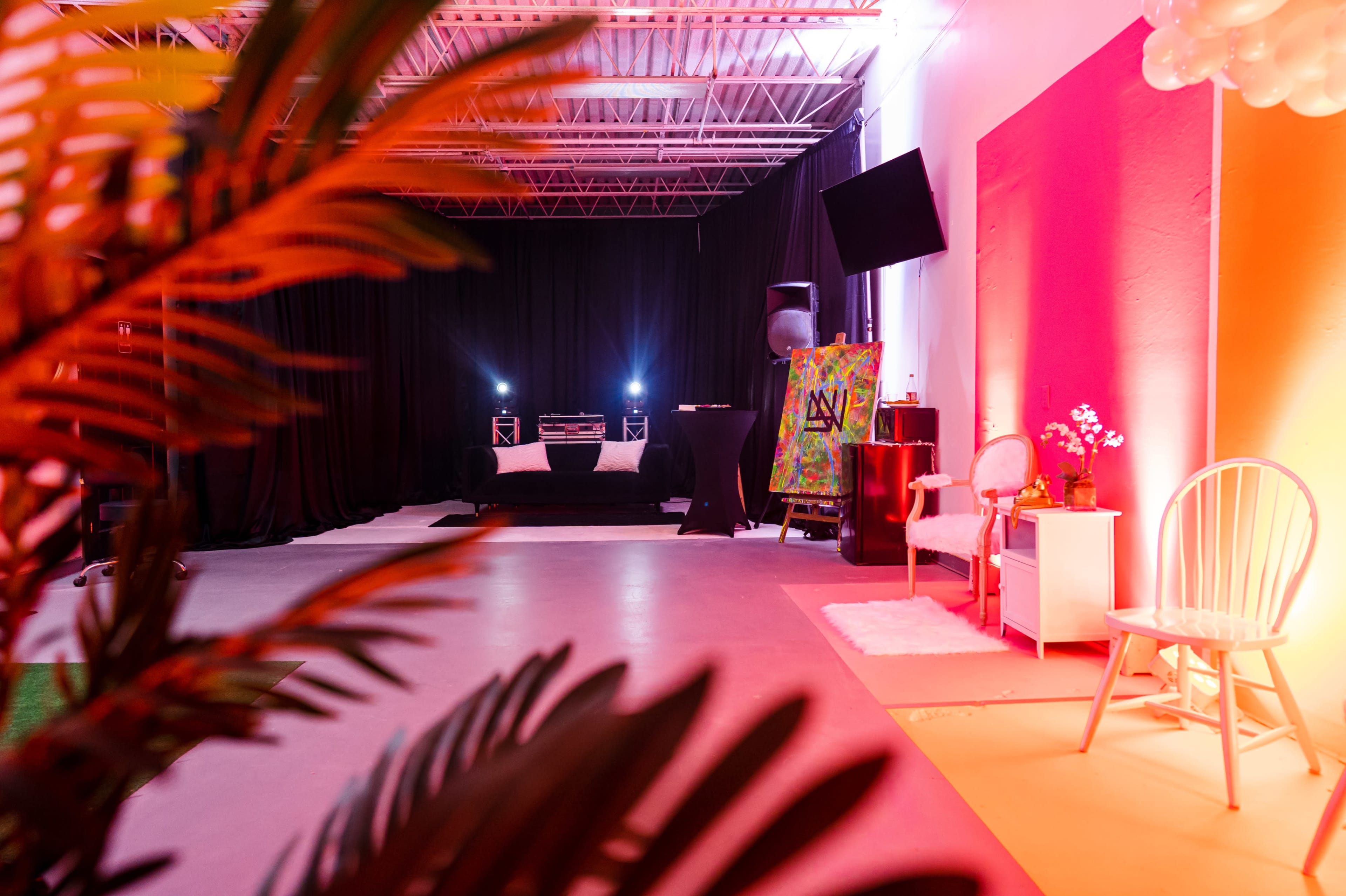 An event space featuring a DJ setup in the background, a decorated table, and a chair with a side table under colorful lighting.