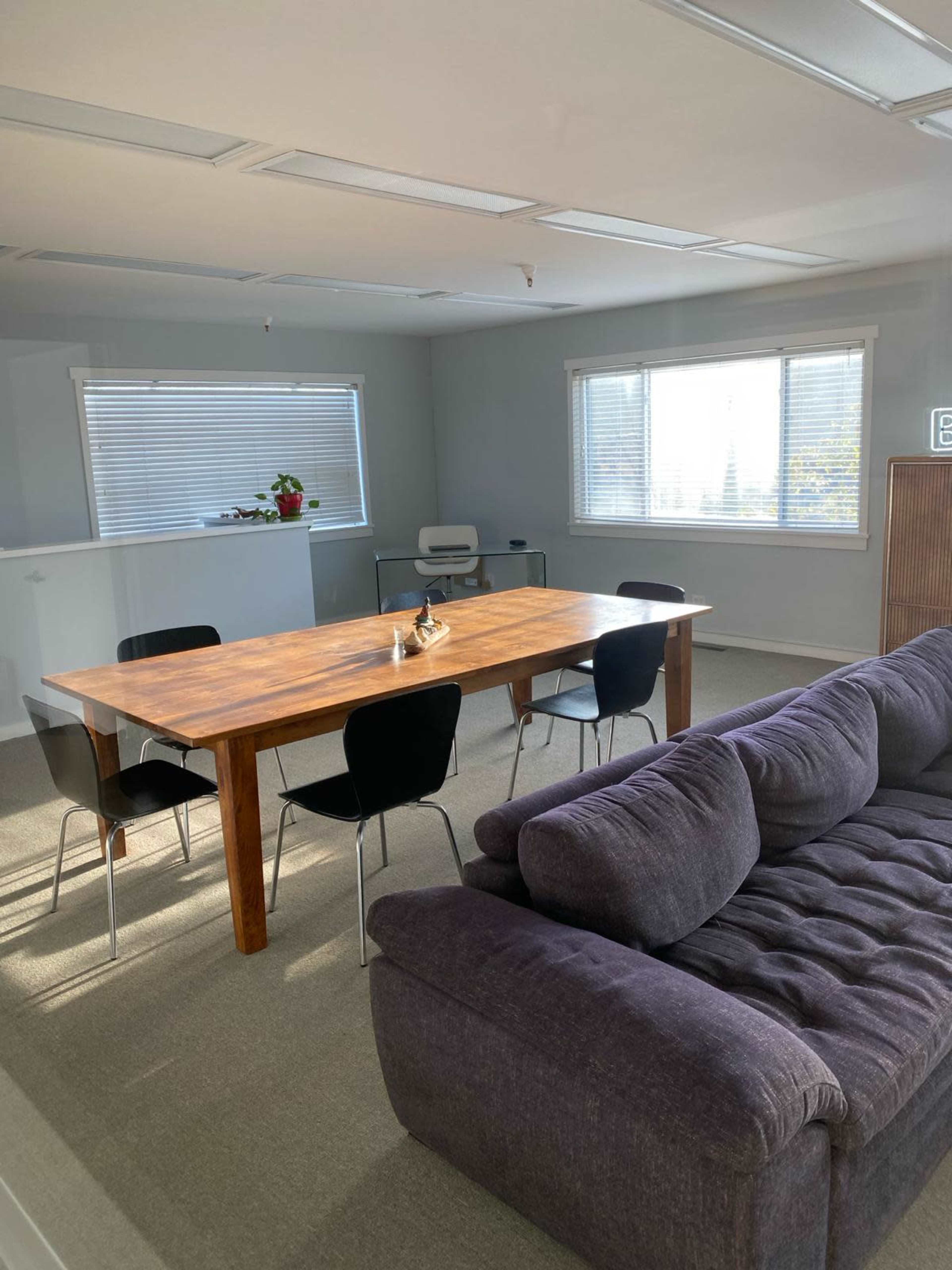 A large wooden table surrounded by black chairs is positioned in a well-lit office space with a gray couch and windows providing natural light.