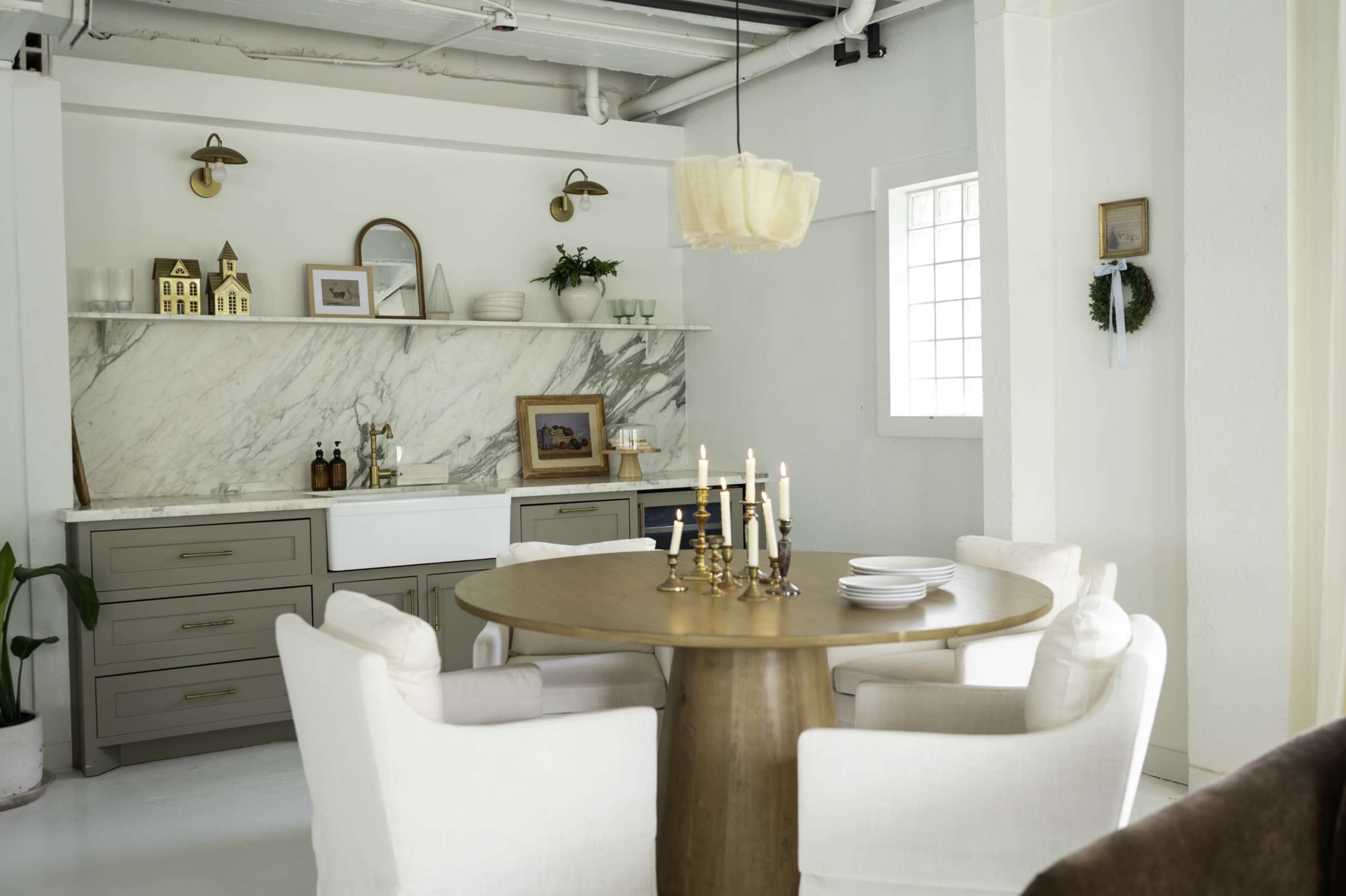 A round wooden dining table is set with plates and candles, surrounded by white upholstered chairs in a bright, minimalist dining area featuring a marble backsplash and shelves with decorative items.