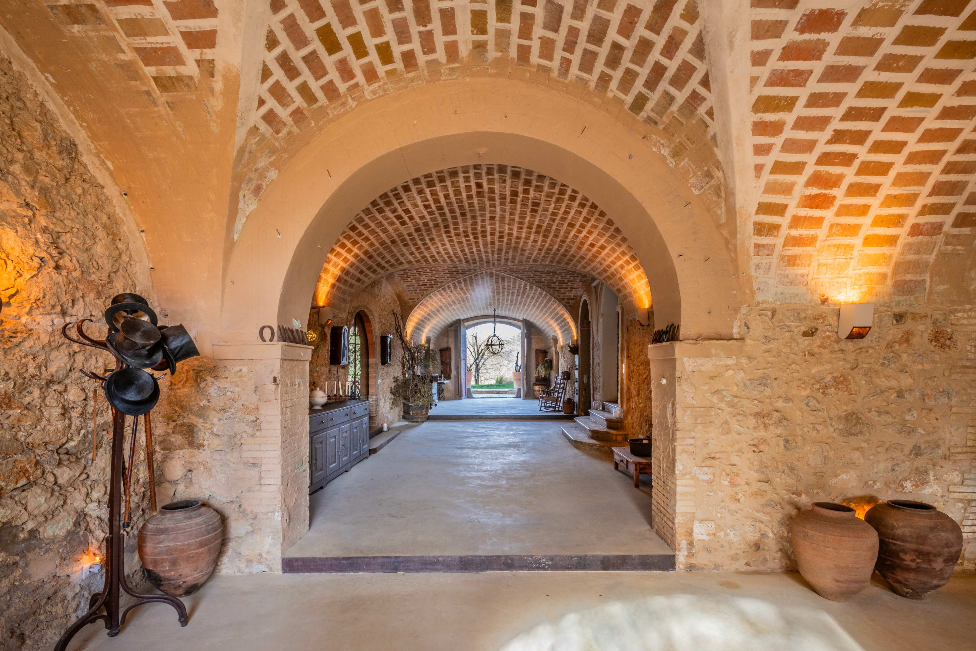 The image features a spacious, arched corridor with brick ceilings and stone walls, leading to an outdoor view framed by double doors.