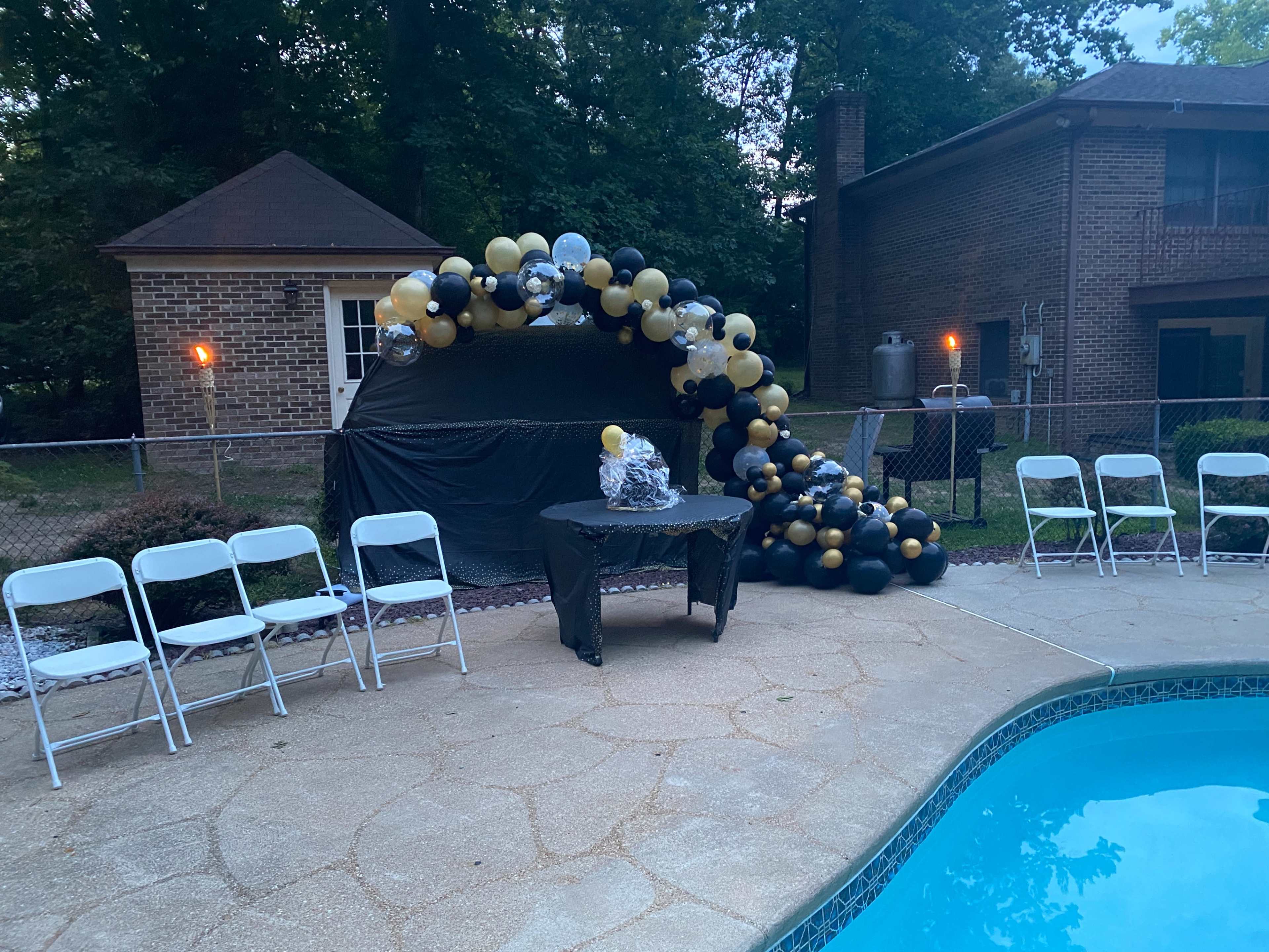 A decorated seating area with a table is set up by a swimming pool, featuring a balloon arch in black and gold colors nearby.