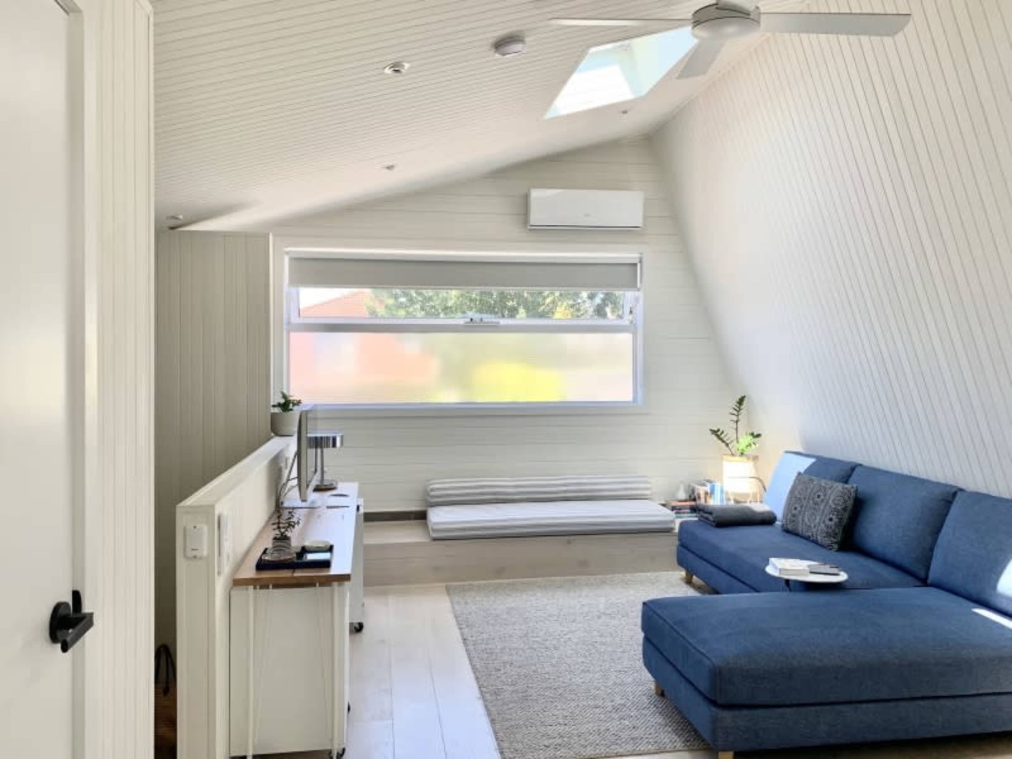 A bright, minimalist living room with a blue couch, a television unit, and a mattress positioned against the wall under a large window.