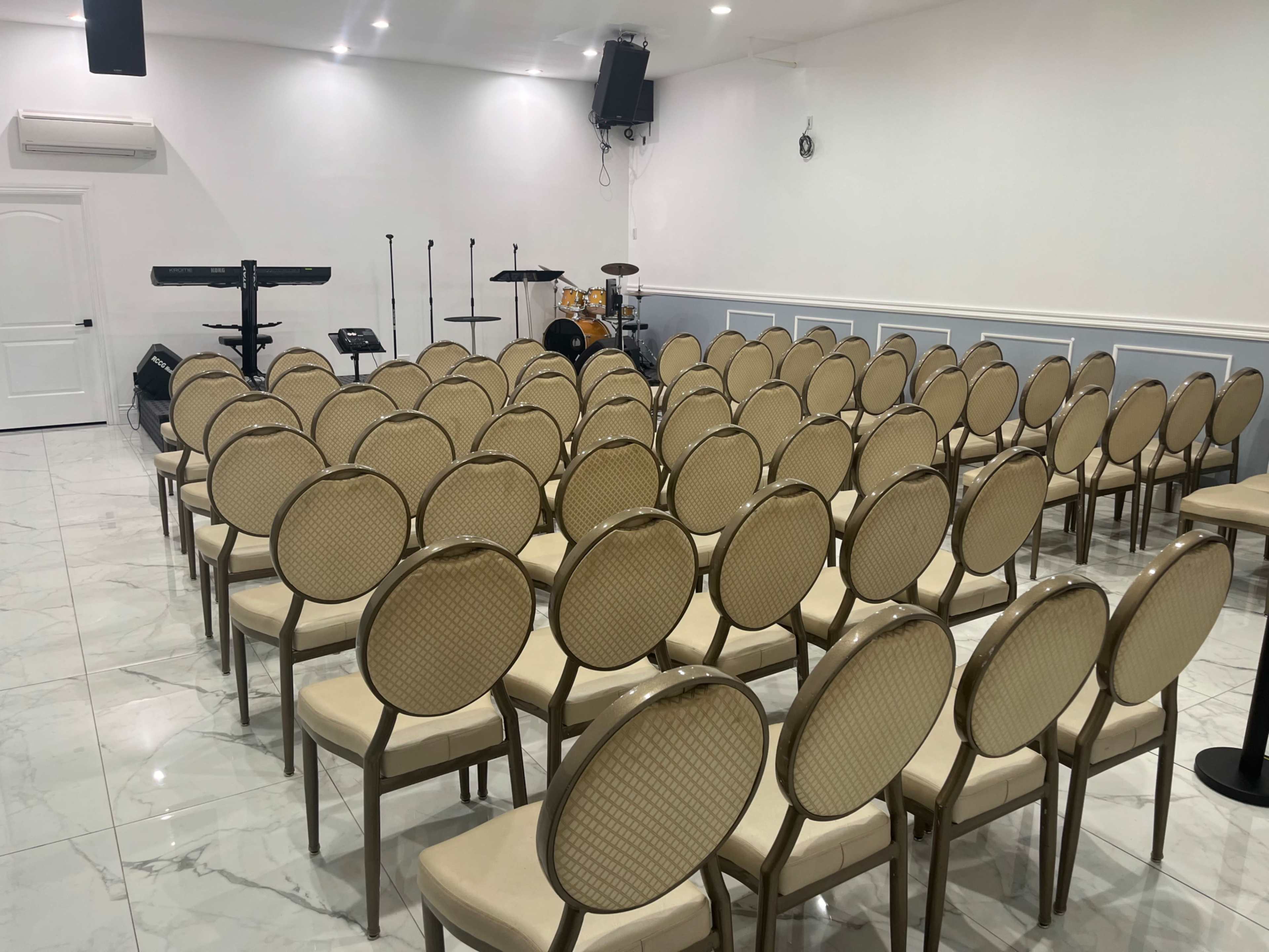 A neatly arranged set of beige chairs facing a small stage equipped with musical instruments in a white-walled room.