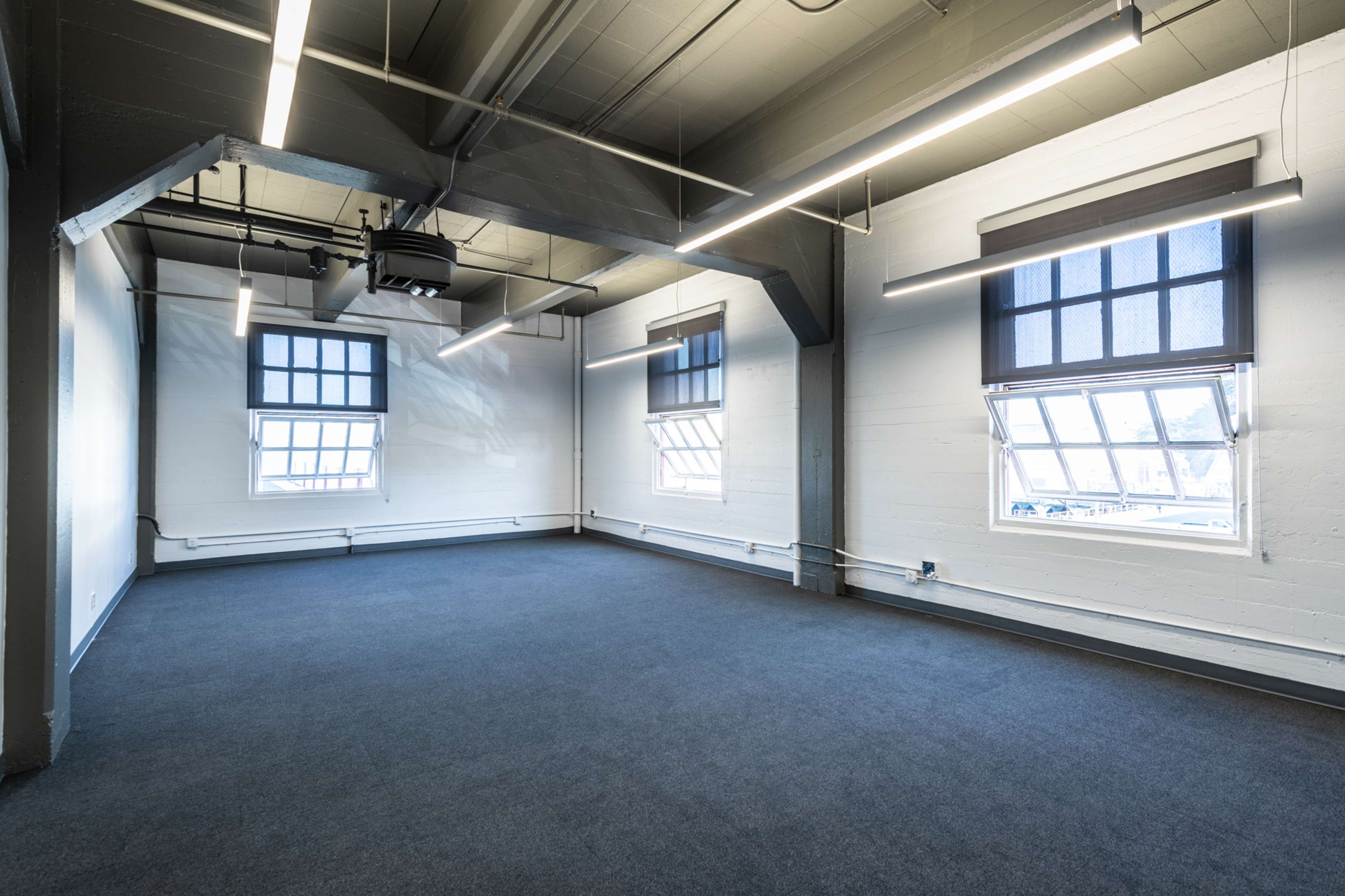 A vacant room with large windows, gray carpet, and modern ceiling lights.